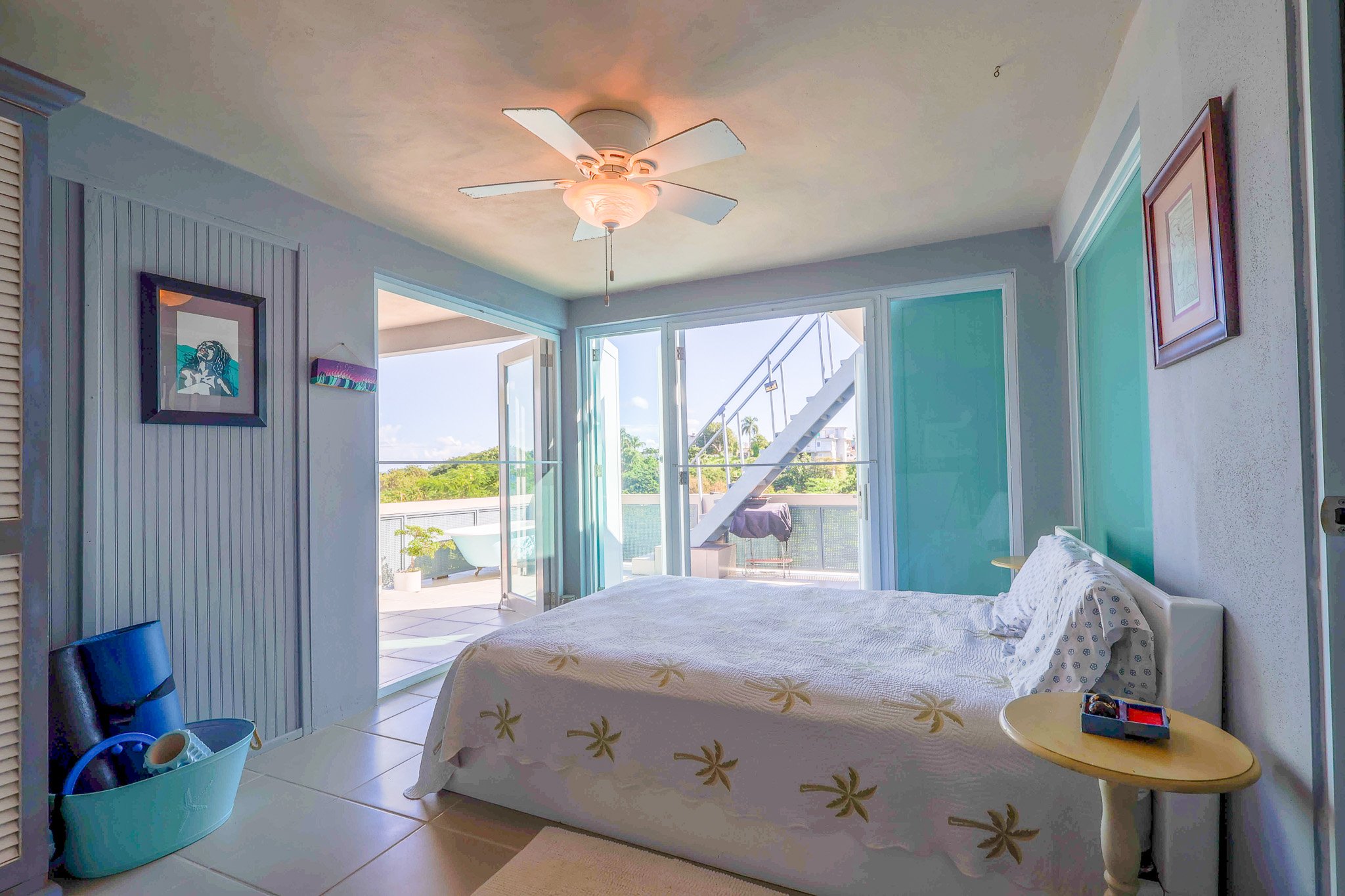 A bedroom with a large glass sliding door leading to a balcony, white walls, a ceiling fan, a bed with white bedding featuring a star and palm tree pattern, and ocean views outside.