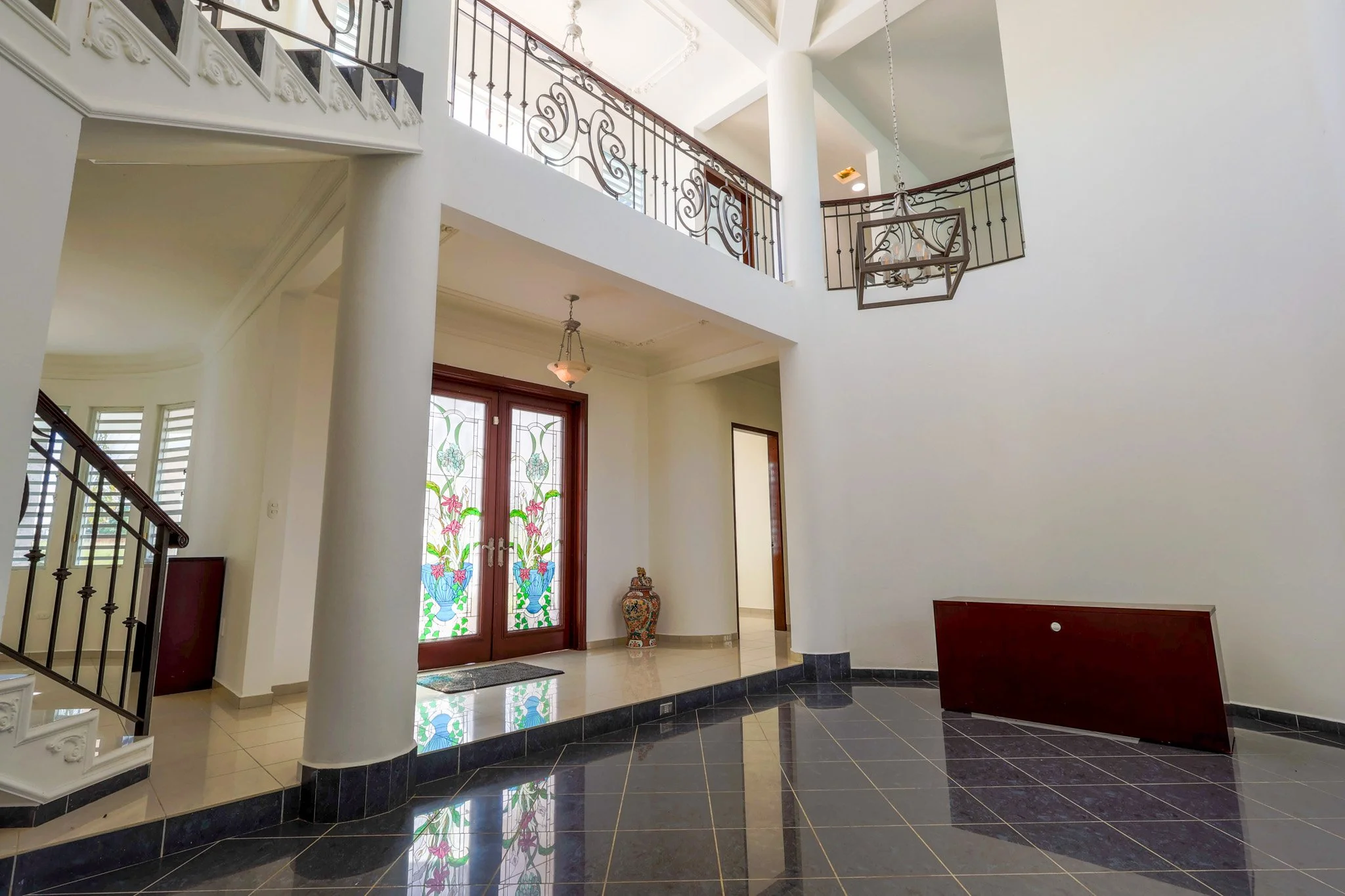 Interior of a spacious house entryway with a high ceiling, stained glass front door, tile and black granite flooring, decorative staircase, and ornate lighting fixtures.
