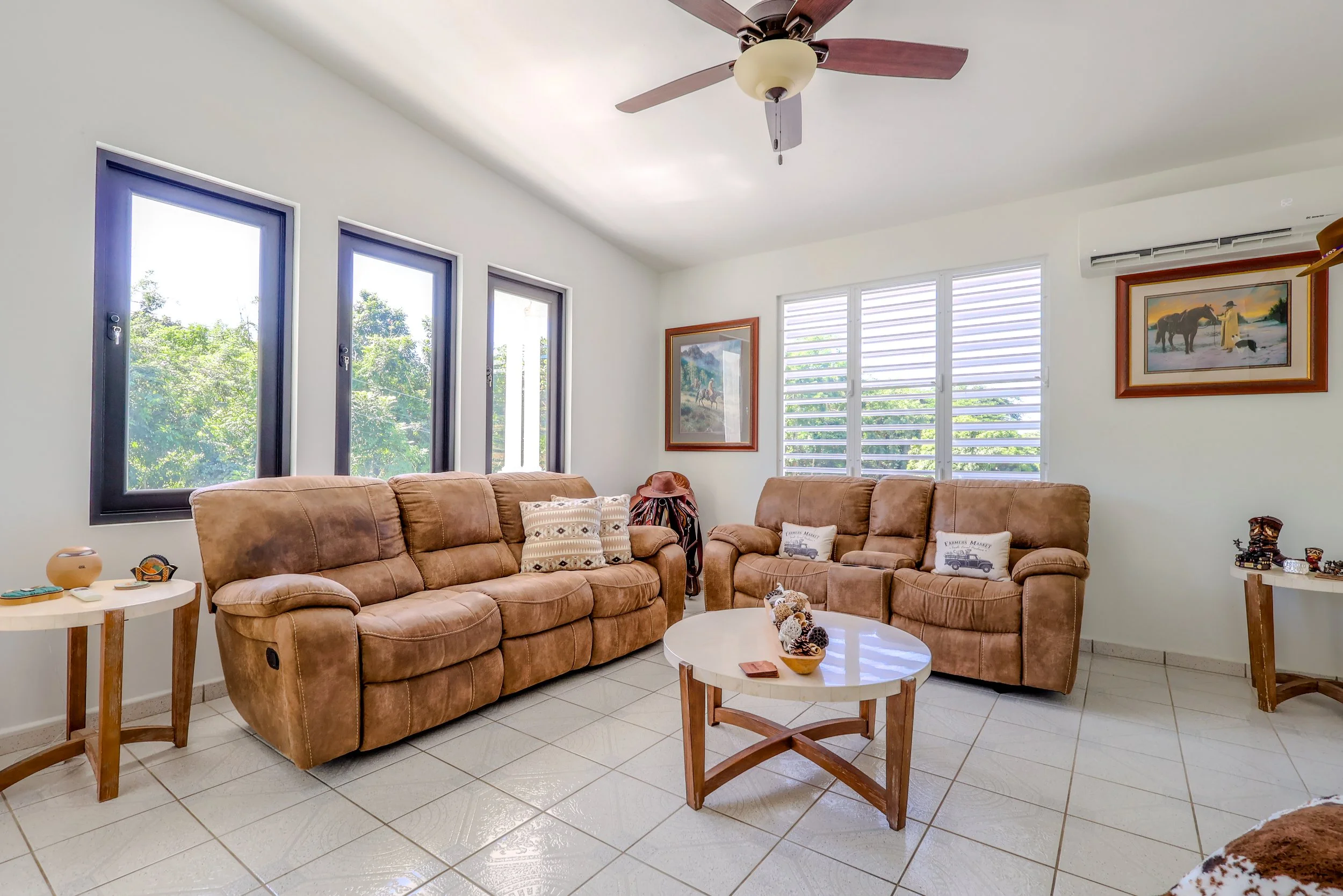 Living room with three brown sofas, white round coffee table, side tables, artwork, ceiling fan, air conditioner, and windows with greenery outside.