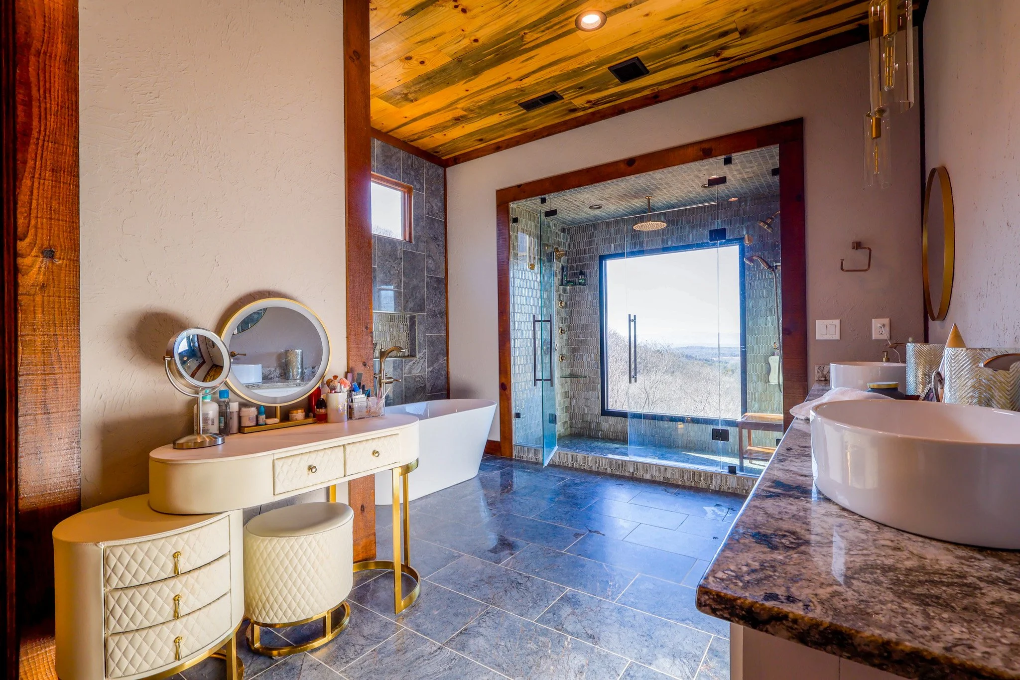 Modern bathroom with wooden ceiling, bathtub, glass-enclosed shower with large window, granite countertop with vessel sinks, and vanity with mirror and personal care items.