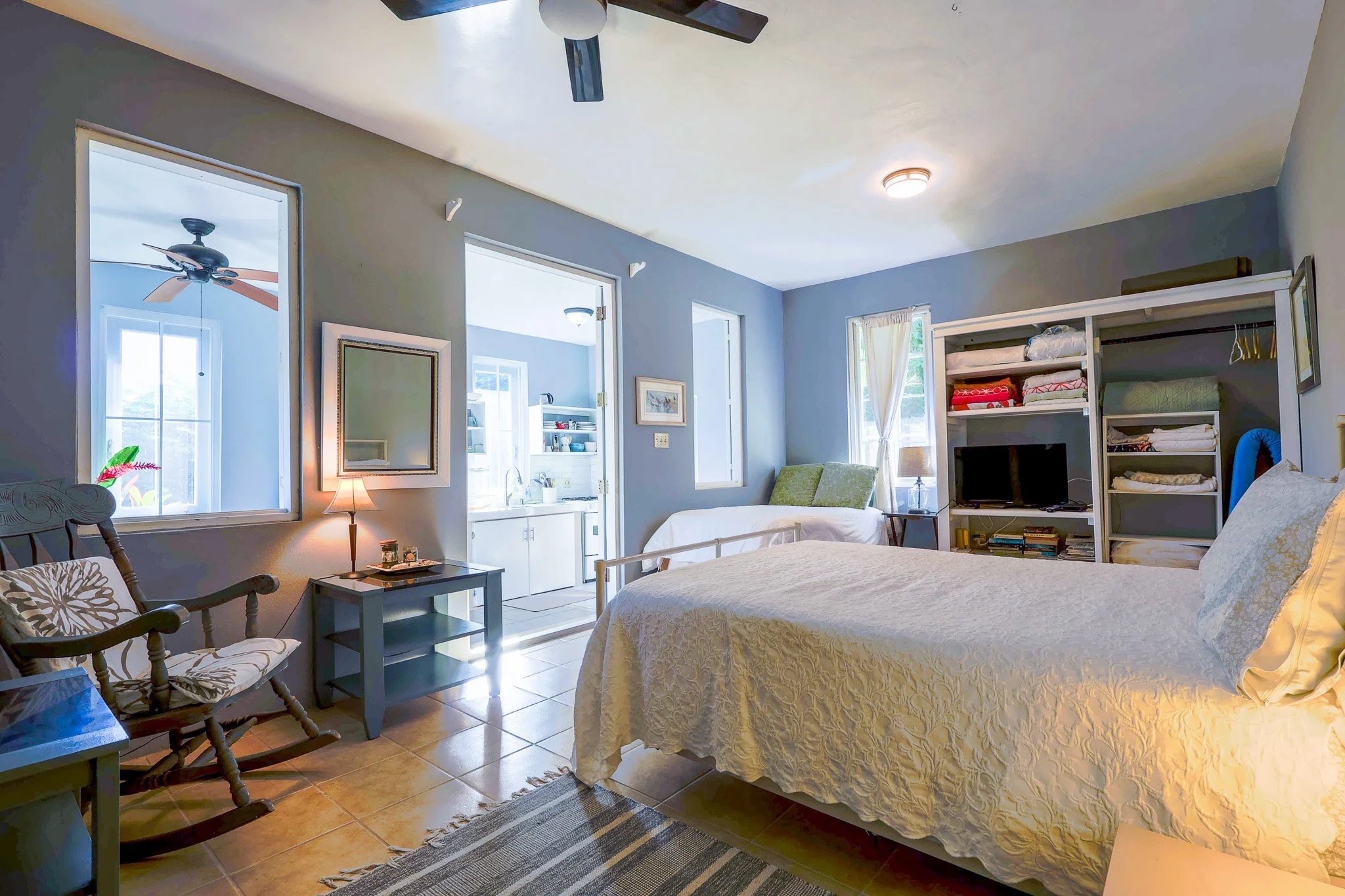 A cozy bedroom with a large bed covered in a cream quilt, a wooden rocking chair, and a nightstand with a lamp. There are multiple windows and a door leading to a bright, open bathroom or laundry room. A wall-mounted mirror, a seating area with a gre