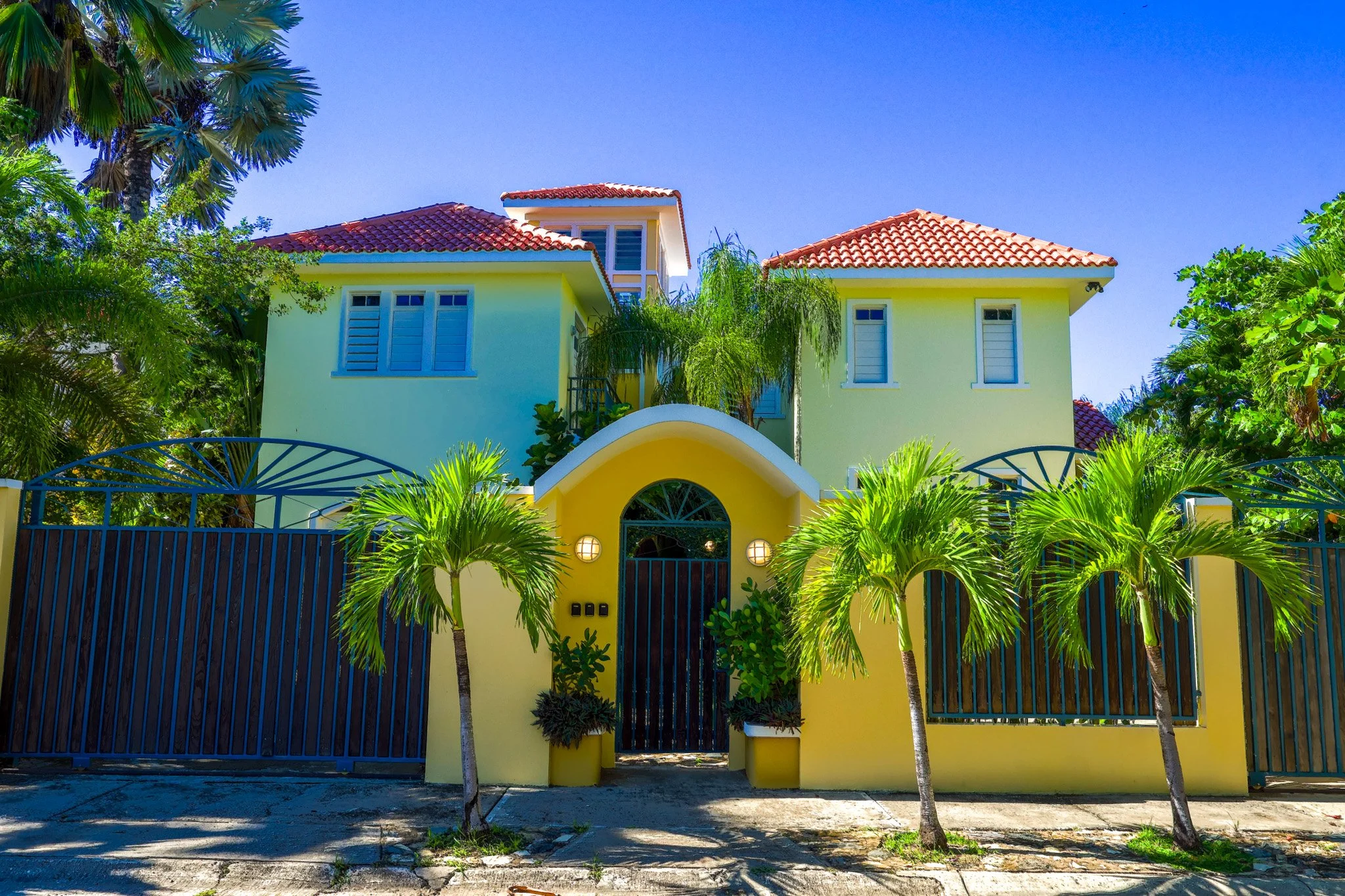 A yellow house with red tiled roof, surrounded by palm trees and greenery, with a black gate and fence, under a clear blue sky.