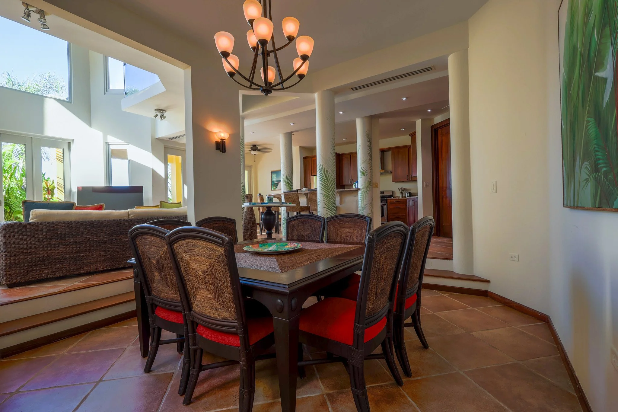 Dining area with a dark wood table, six chairs with red cushions, a chandelier, and a view of an open living room and kitchen with wooden cabinets.
