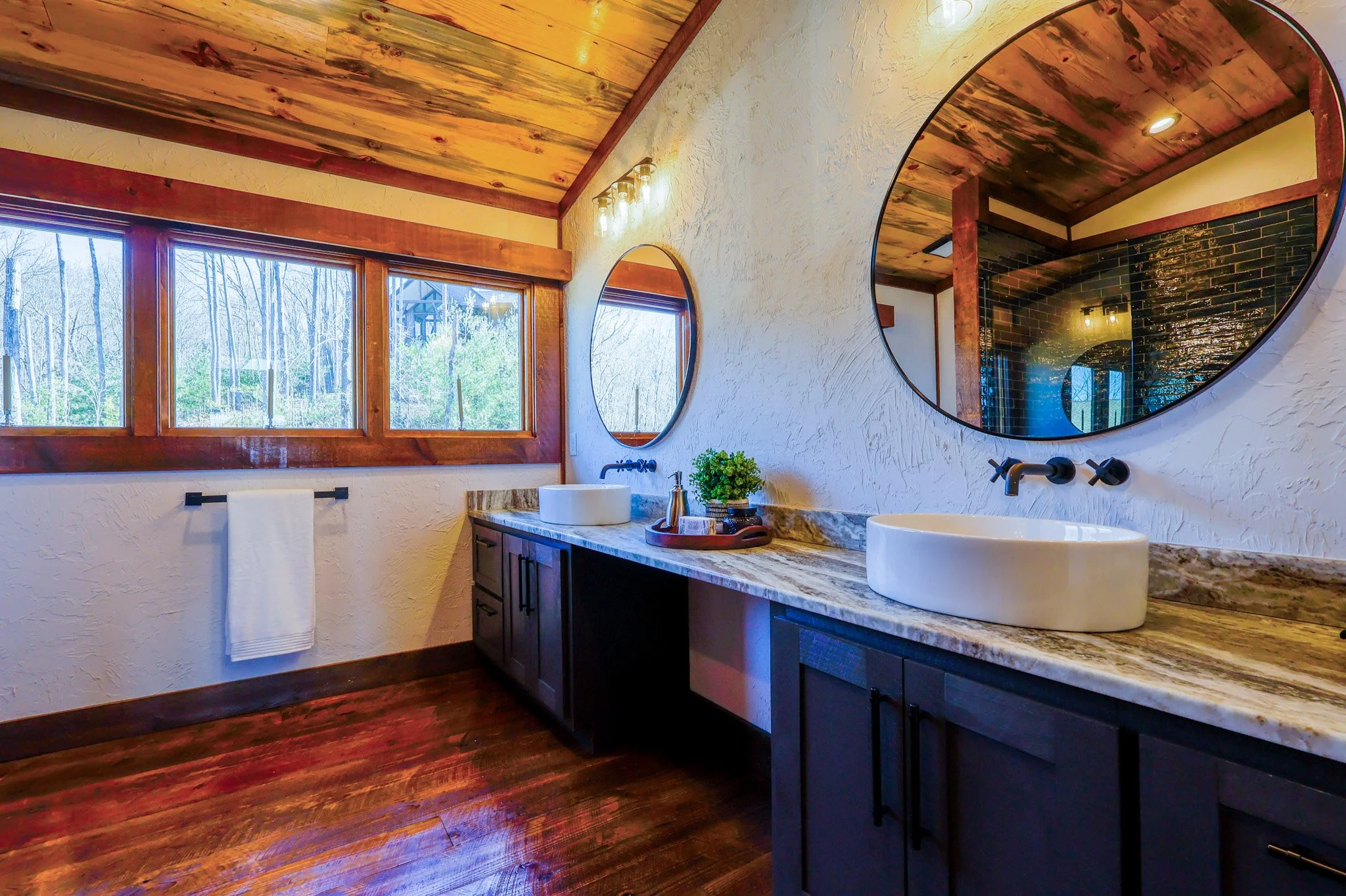 A spacious bathroom with wooden flooring and a matching wooden ceiling, three large windows showing trees outside, white textured walls, two oval mirrors above a dark wooden vanity with a marble countertop, two vessel sinks, black wall-mounted faucet