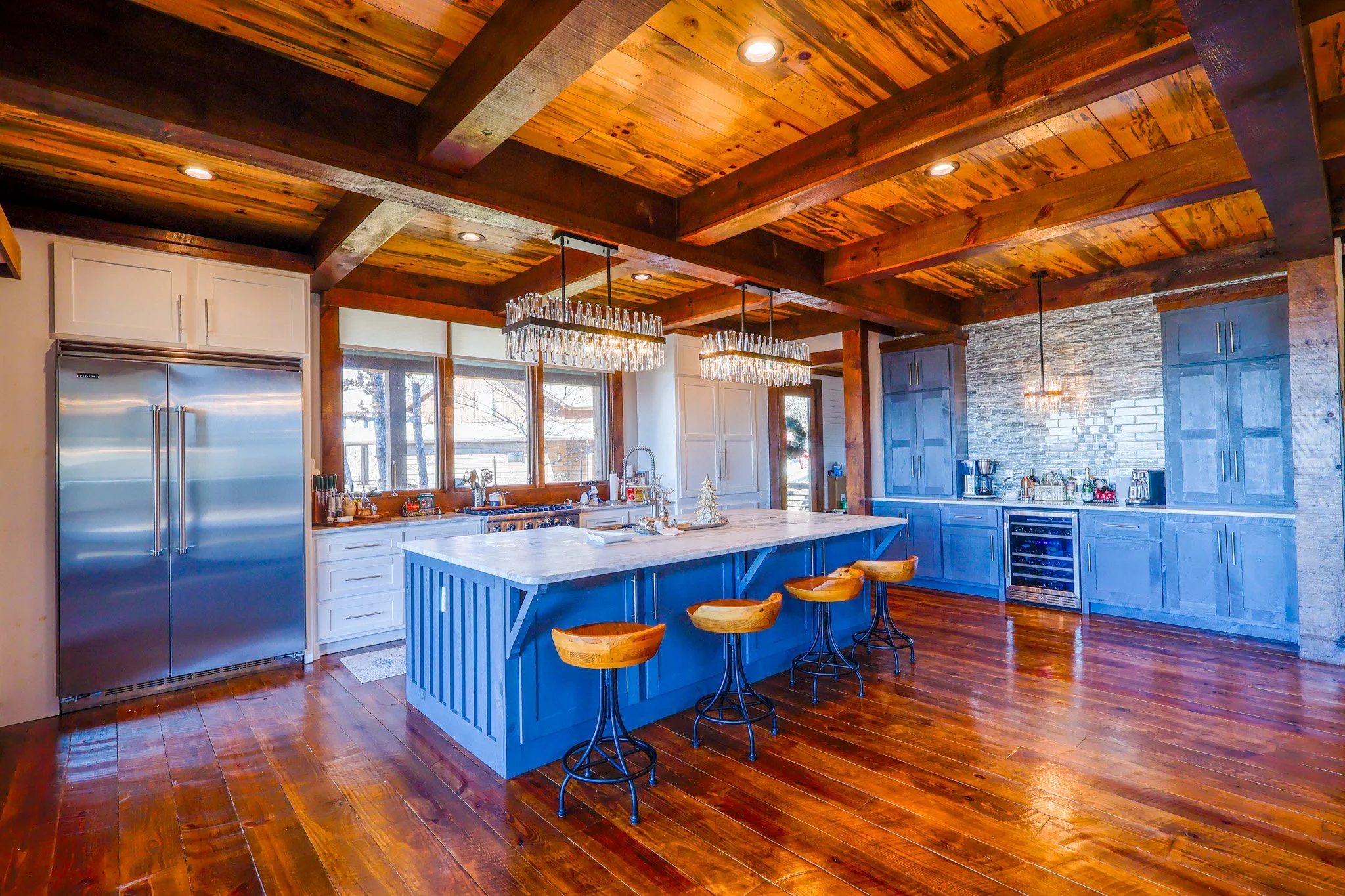 Modern kitchen with wooden ceiling and floors, blue island with bar stools, stainless steel refrigerator, large windows, and stone accent wall.