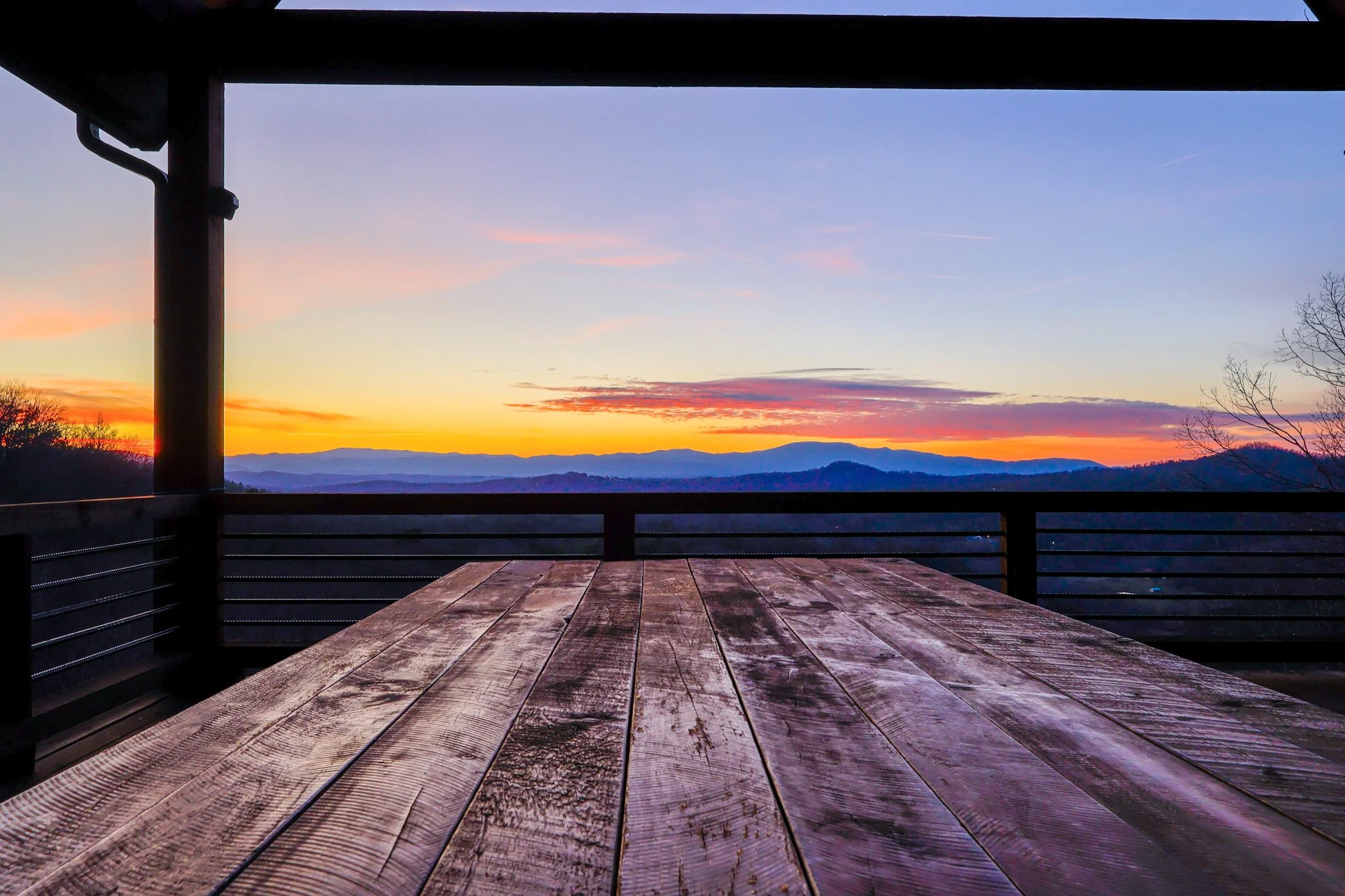 Sunset over mountains viewed from a wooden deck framed by a railing and roof, with a blurry silhouette of trees on the right.