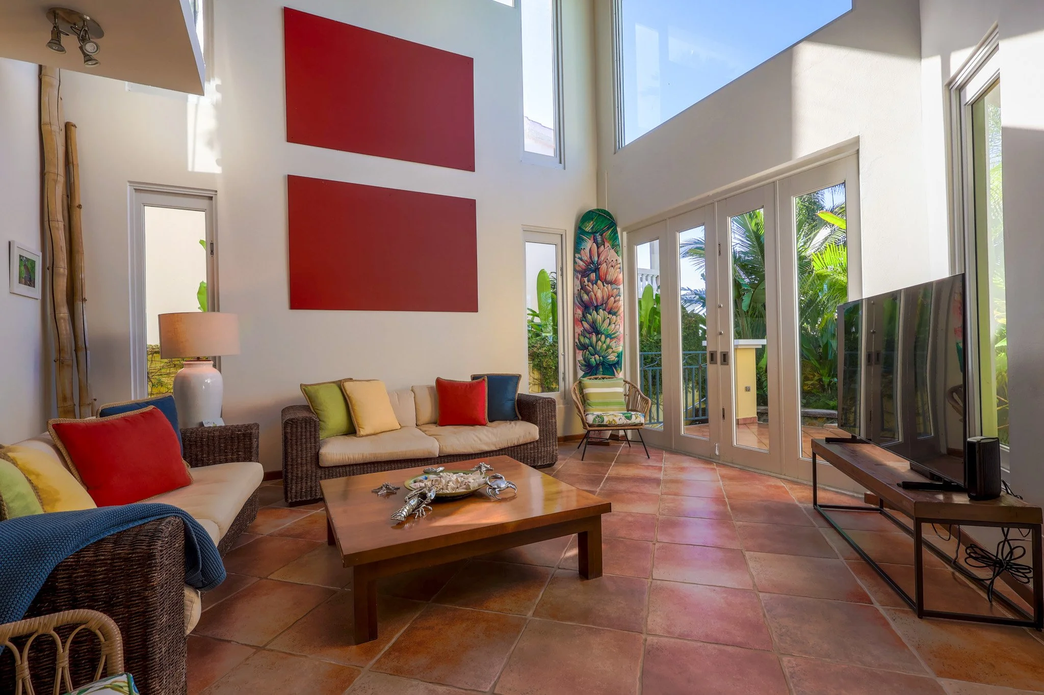 Bright living room with large windows, a beige sofa with colorful pillows, a wooden coffee table, a flat-screen TV on a stand, and tropical plants outside.