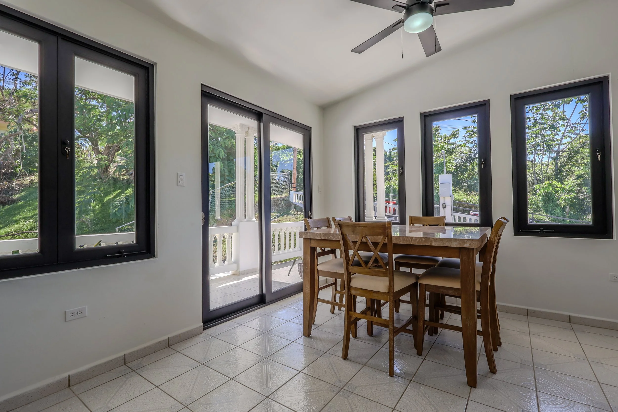 A dining room with five windows and a sliding glass door, a ceiling fan with a light, a wooden dining table, and six chairs, with a view of a green garden outside.