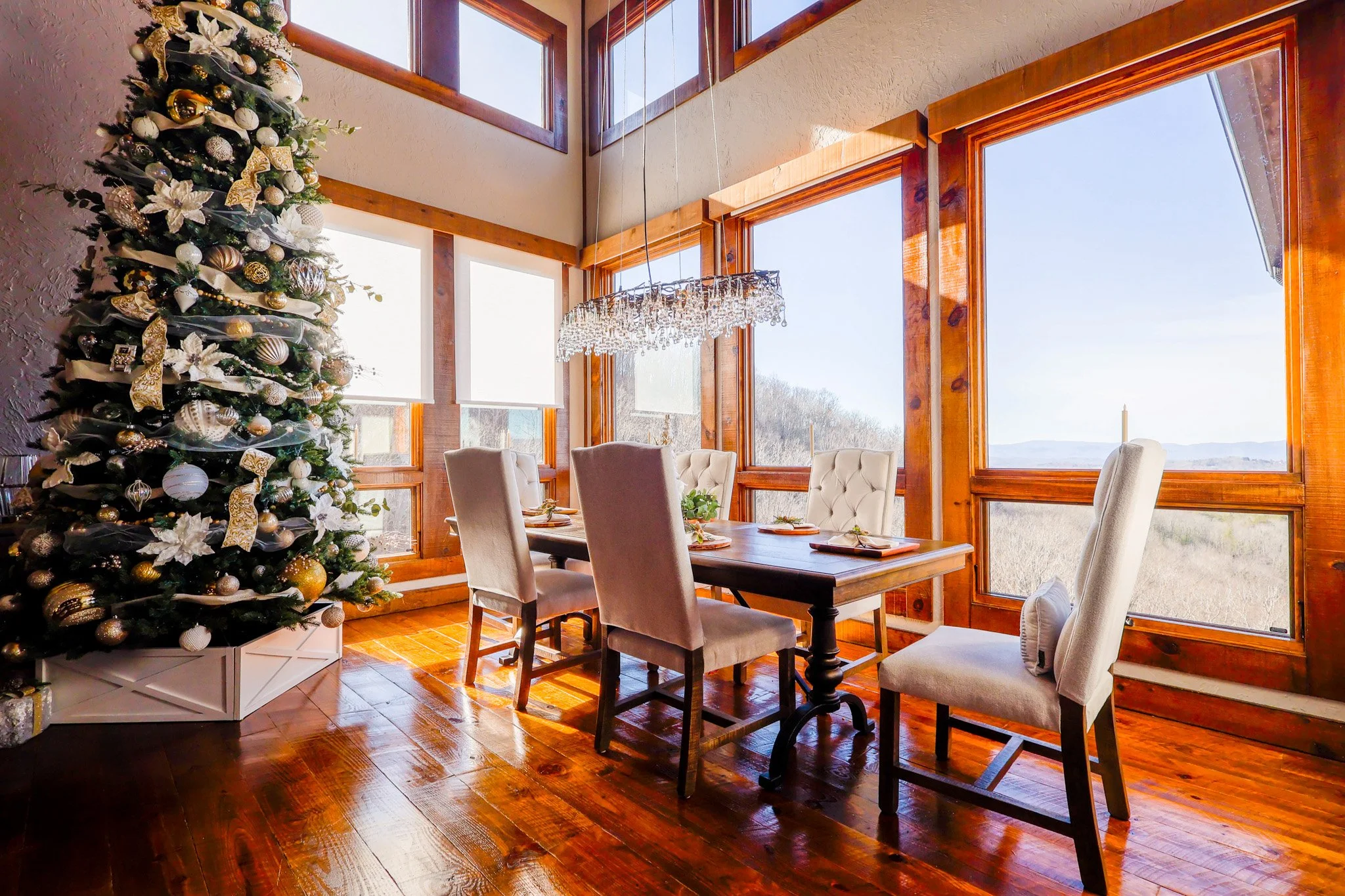 A dining room with a decorated Christmas tree, a wooden dining table with chairs, large windows showing a landscape outside, and a chandelier.