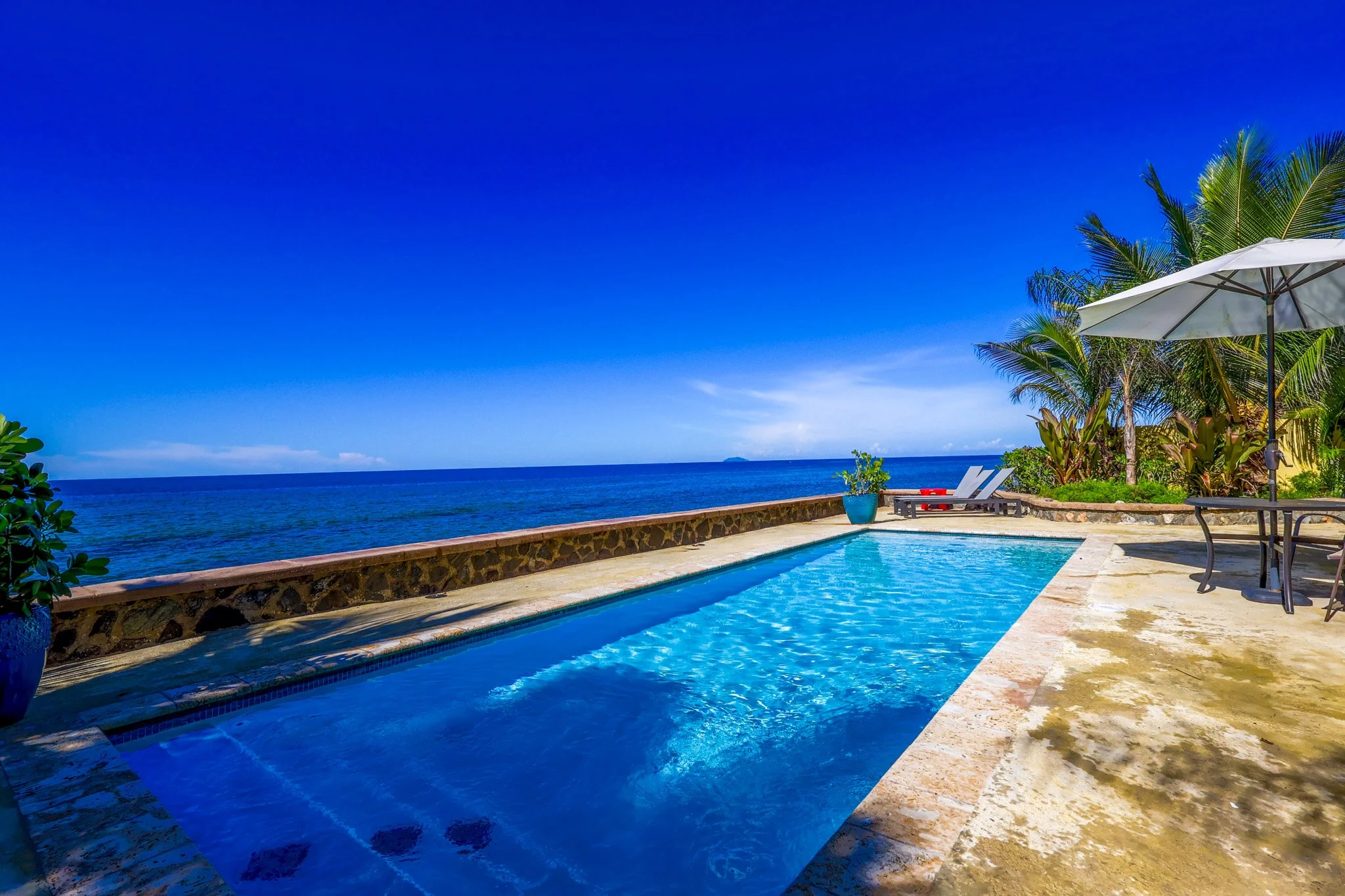 A seaside poolside area with two lounge chairs under an umbrella, surrounded by palm trees, overlooking the ocean and a clear blue sky.