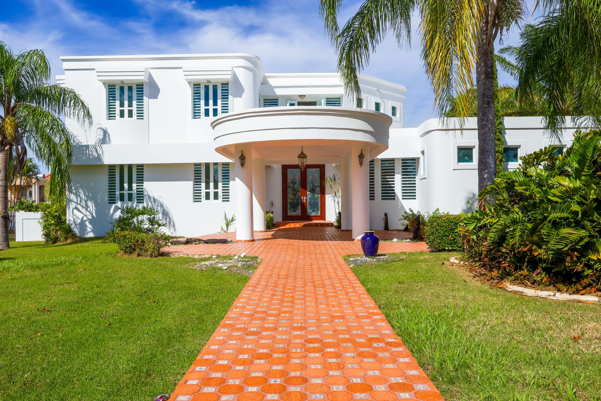 White modern house with curved entrance, surrounded by palm trees, green lawn, and a brick walkway leading to the front door, under a partly cloudy sky.