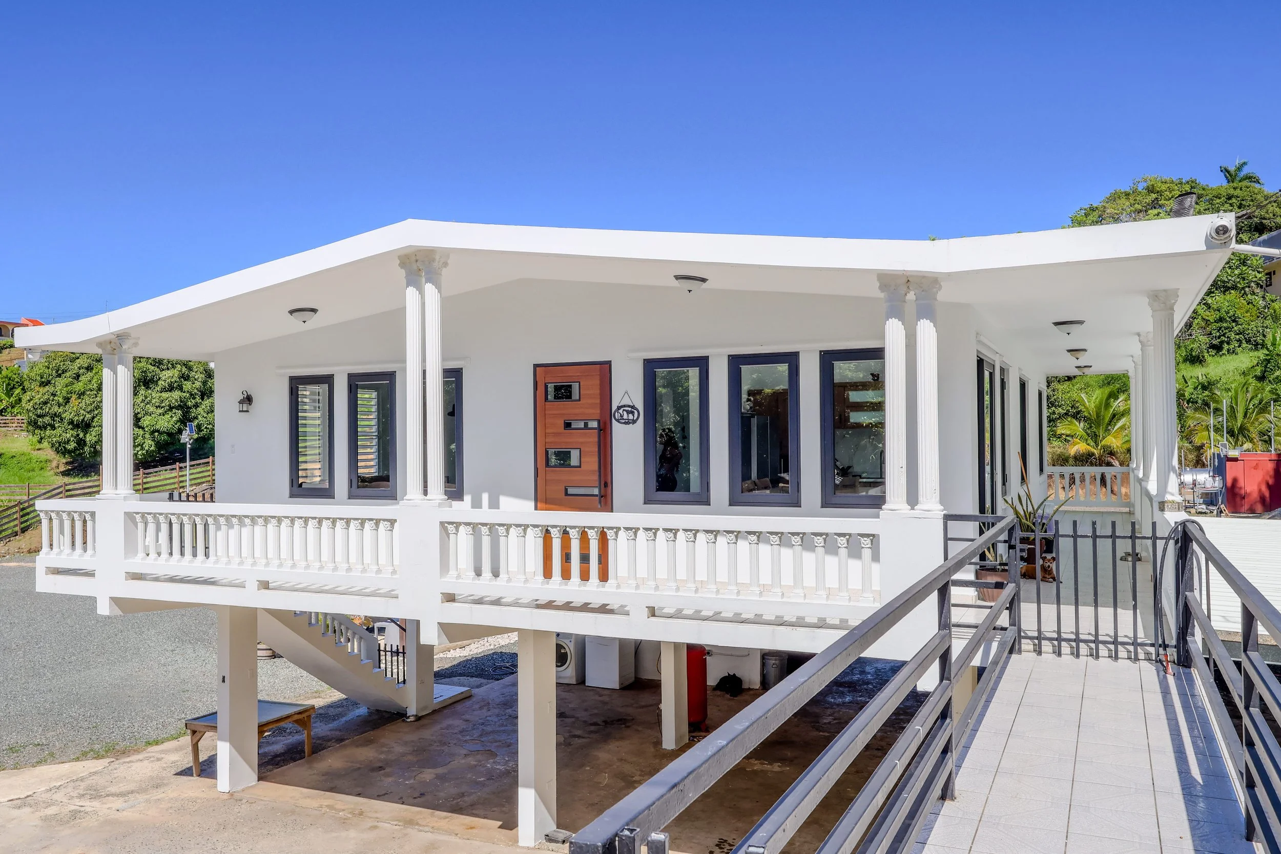 White modern house with a balcony, multiple windows, and a unique curved roof, set against a bright blue sky and green foliage.