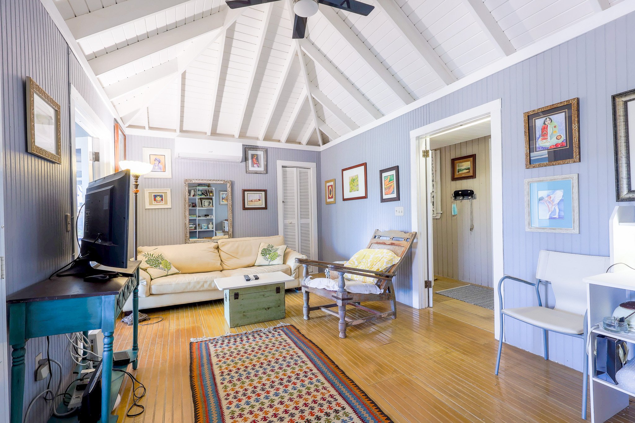 Living room with light wood floor, blue paneled walls, white vaulted ceiling, beige sofa, wooden chair with yellow cushion, patterned rug, TV on teal stand, and framed artwork on walls.