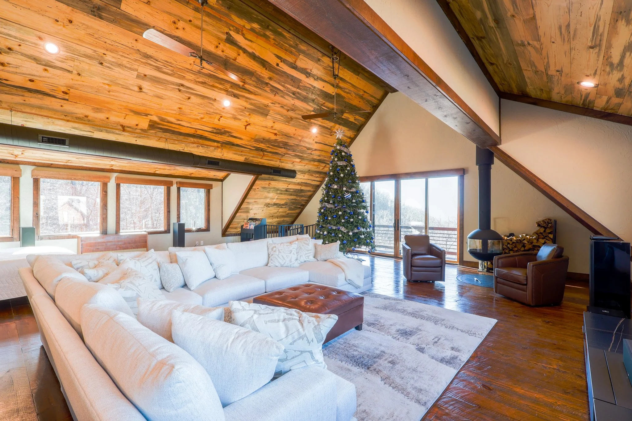 Living room decorated for Christmas with a large Christmas tree, white sectional sofa, leather armchairs, wood stove, and large windows with view of outdoors, featuring wood-paneled ceiling and hardwood floors.