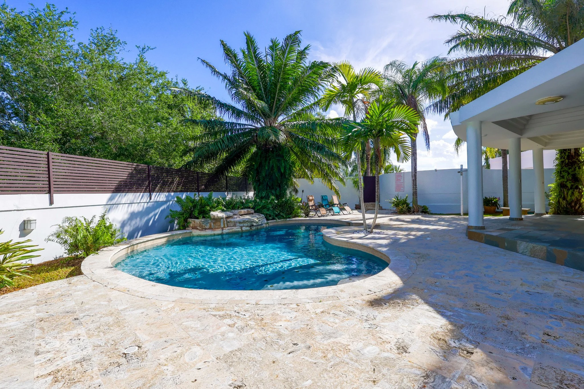 Swimming pool surrounded by lounge chairs, palm trees, and a shaded patio area with columns, in a tropical backyard setting with clear blue sky.