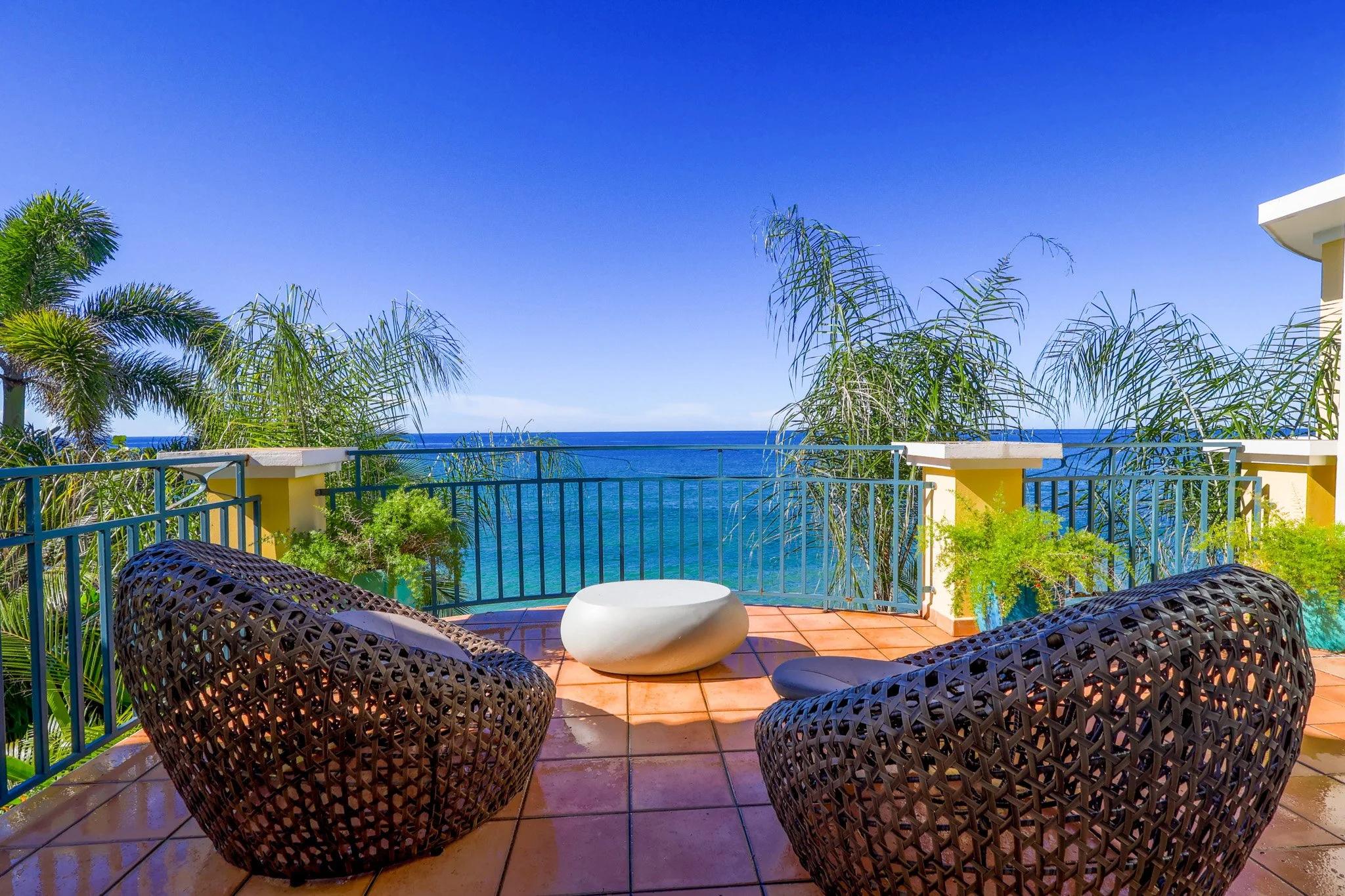 Balcony overlooking the ocean with two wicker chairs, a white stone table, green plants, palm trees, and a blue sky.