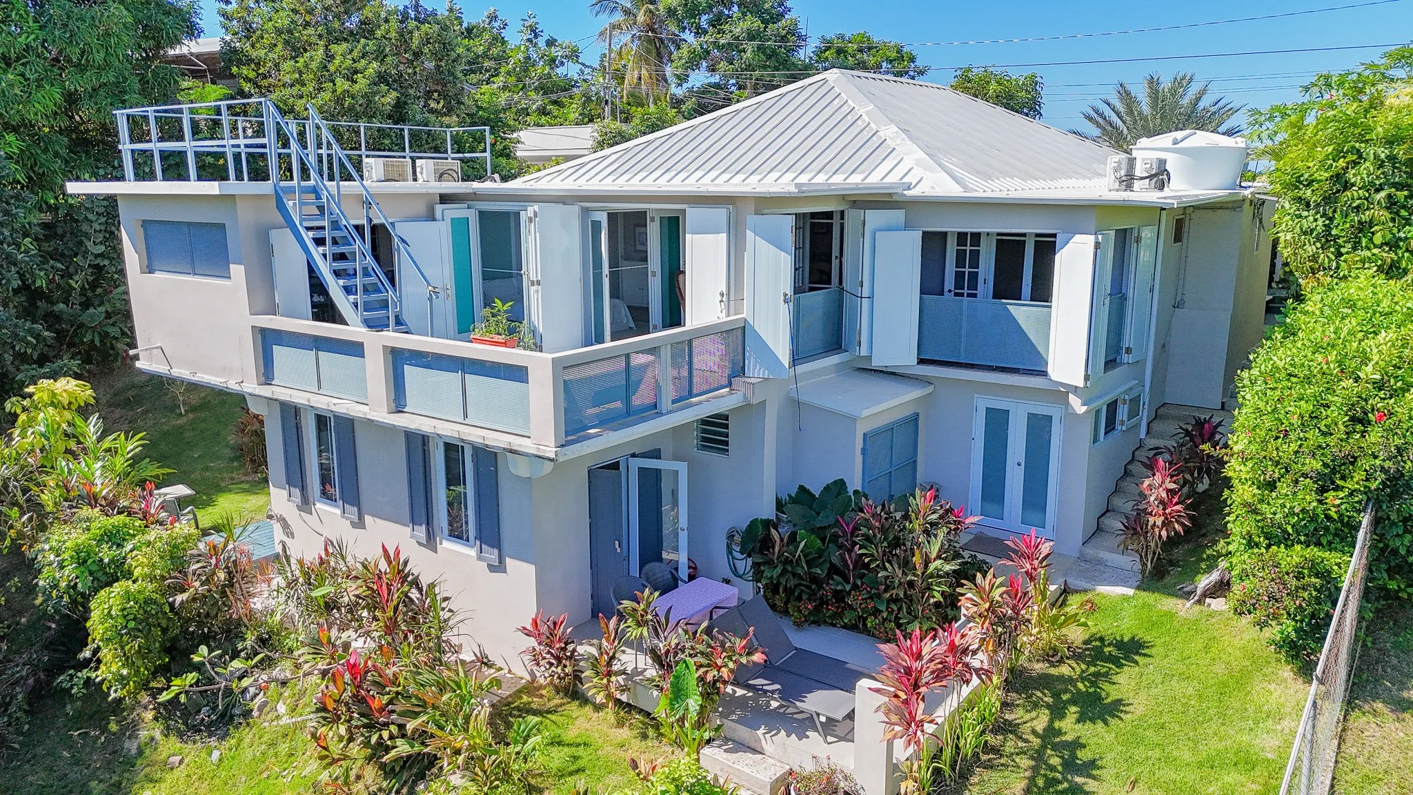 A modern two-story house surrounded by lush green plants and trees. The house has a white exterior with multiple balconies and open doors, a metal roof, and a rooftop terrace with a blue railing. There are outdoor lounge chairs and a table on the gro