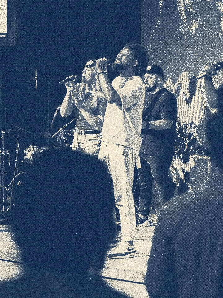 A live music performance with a singer or musician wearing a short-sleeved shirt and jeans, singing or speaking into a microphone. Several other band members or performers are visible in the background on a stage with lighting and equipment.
