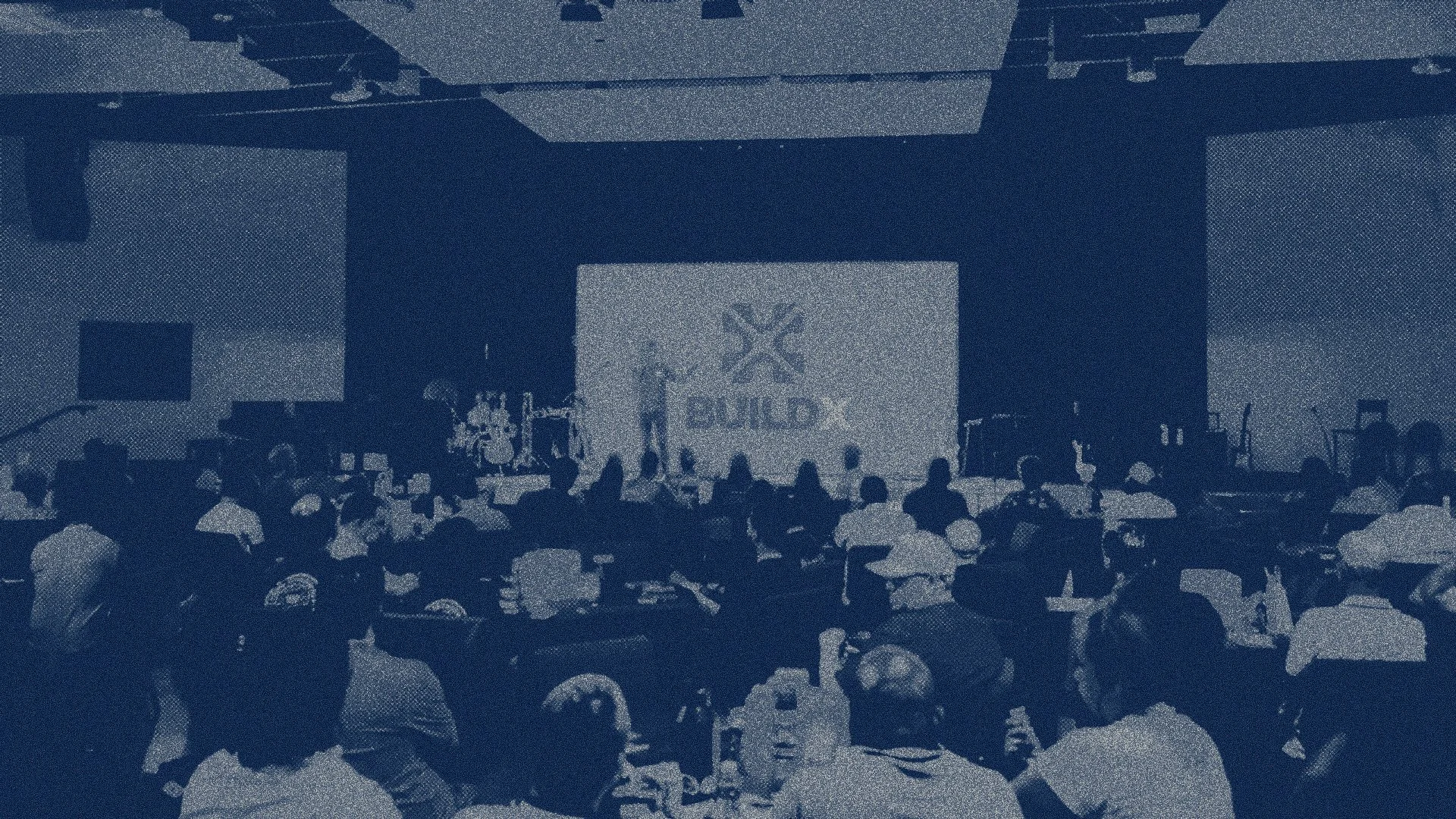Large conference room filled with seated attendees facing a stage with a large screen displaying the word 'BUILD' and a logo. The stage has musical instruments and speakers, suggesting a conference or presentation event.
