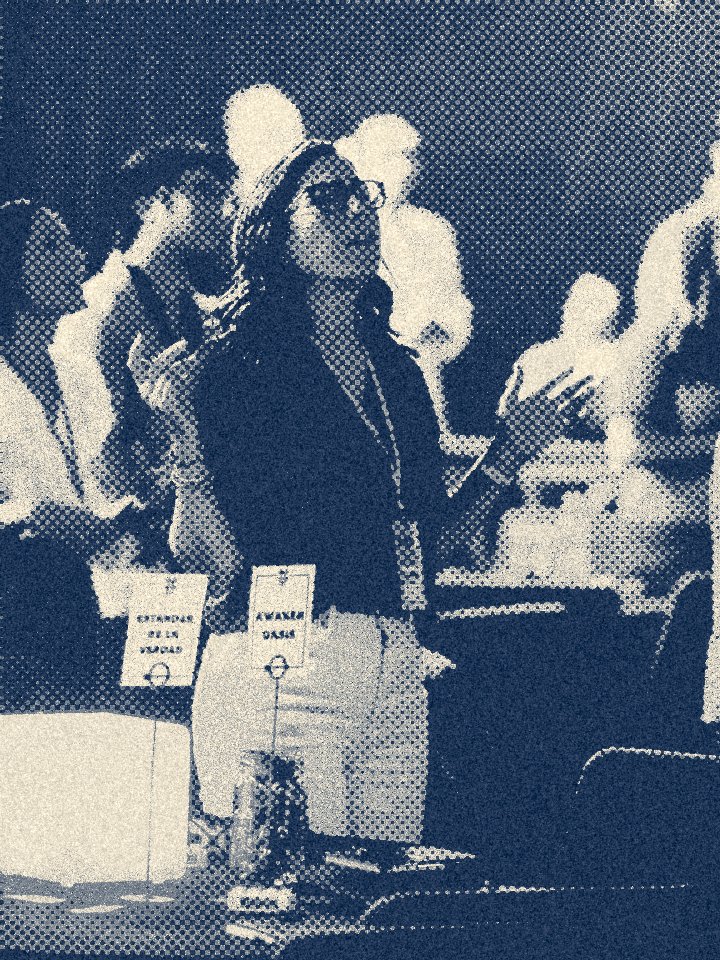 A woman with sunglasses and a hat at an indoor event, standing near a table with signs and a packed crowd in the background.