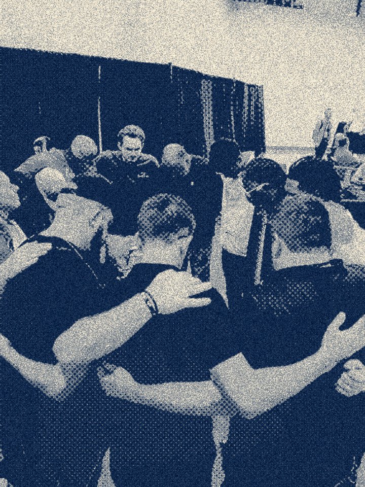 Several people with their heads bowed and arms around each other's shoulders, gathered in a circle in a room at a meeting or event.