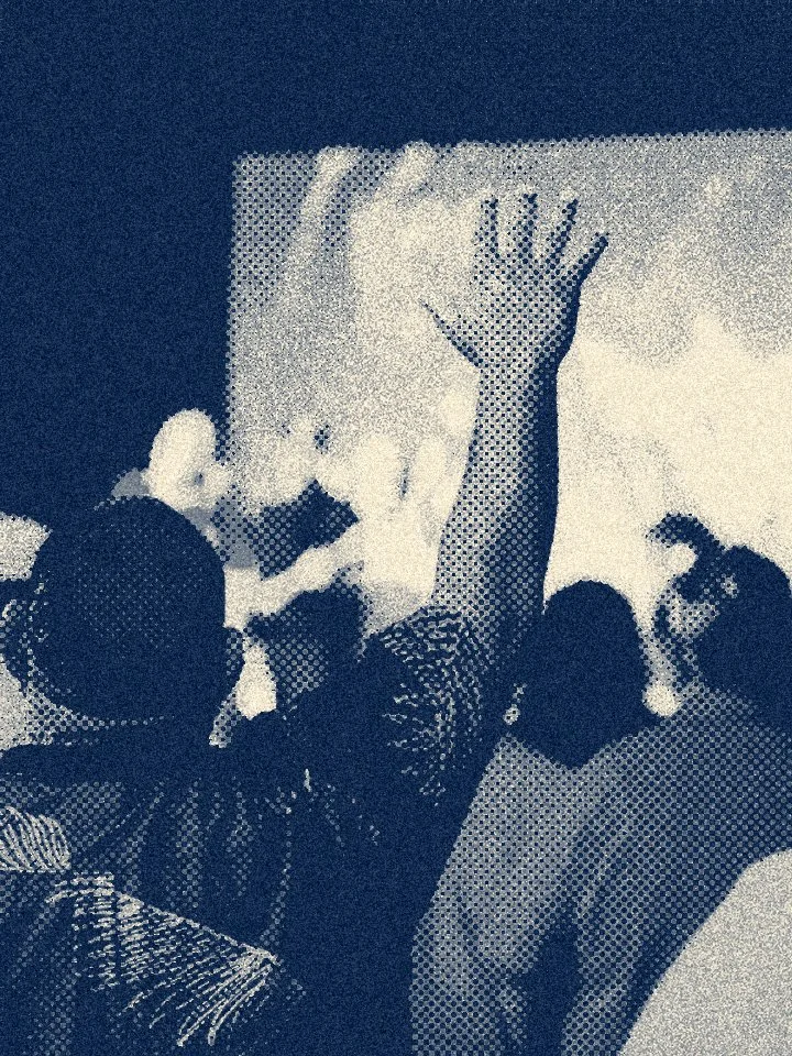 A person in a patterned shirt raises their hand in a crowd at a gathering or event, with others around them and a screen or projection in the background.
