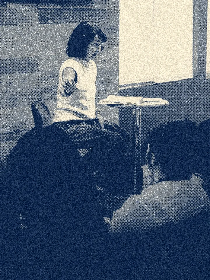 A young girl gestures with her hand while standing next to an adult at a table in an indoor setting.