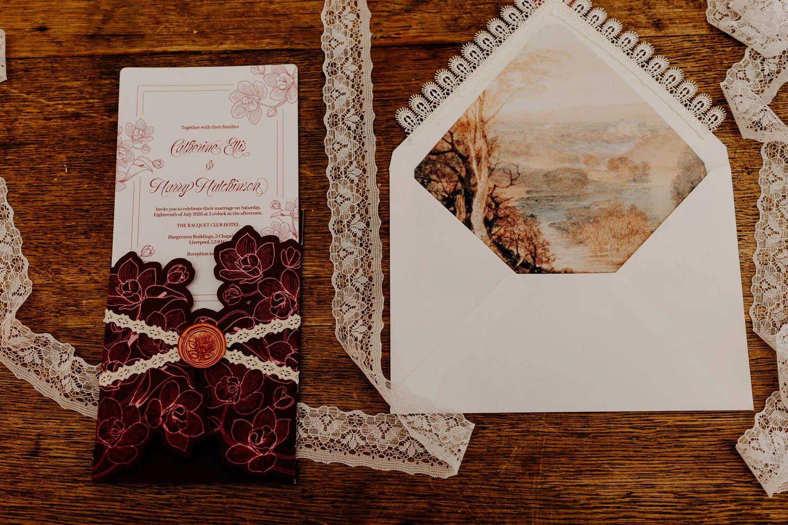 Wedding invitation on a wooden surface with lace borders, featuring a floral design and a red wax seal, alongside an open envelope with a landscape painting inside.
