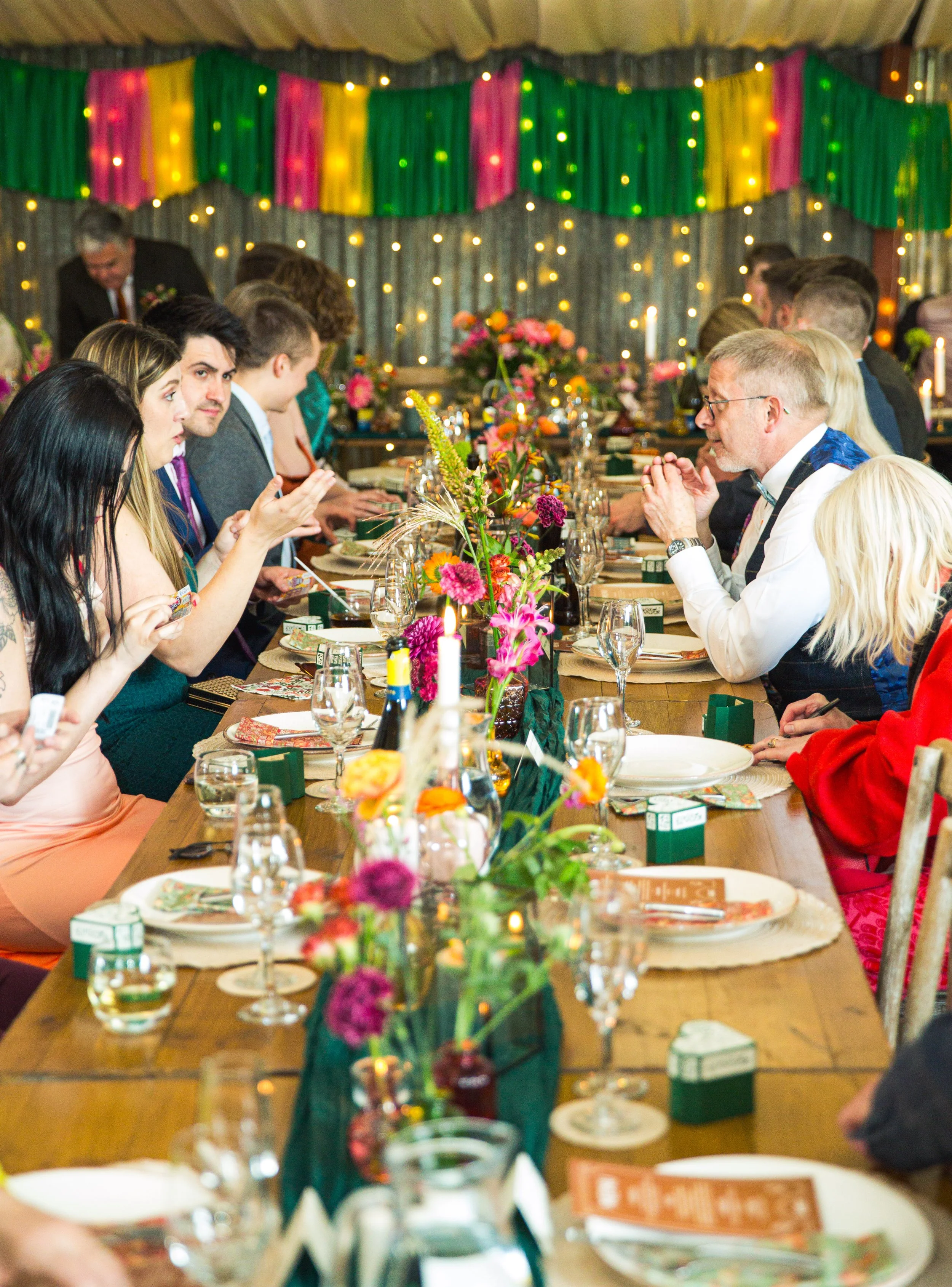 People sitting at a long decorated table with colorful flowers, candles, and tableware, attending a festive celebration under a tent with colorful banners and string lights.
