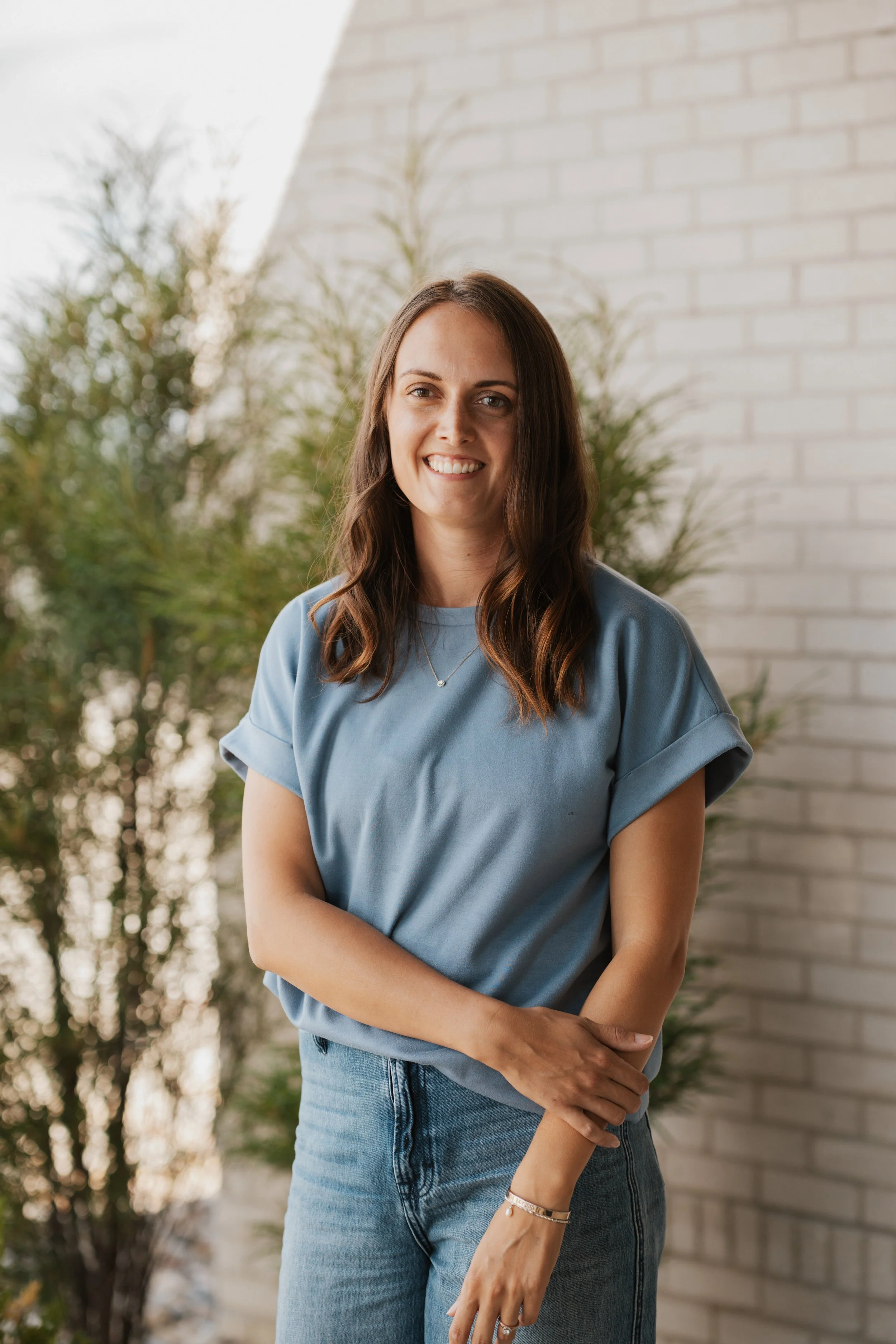 Marquel Stuedemann, Christian writer and author, with long brown hair, smiling, is wearing a light blue t-shirt and jeans, standing outdoors near a white brick wall with greenery in the background.