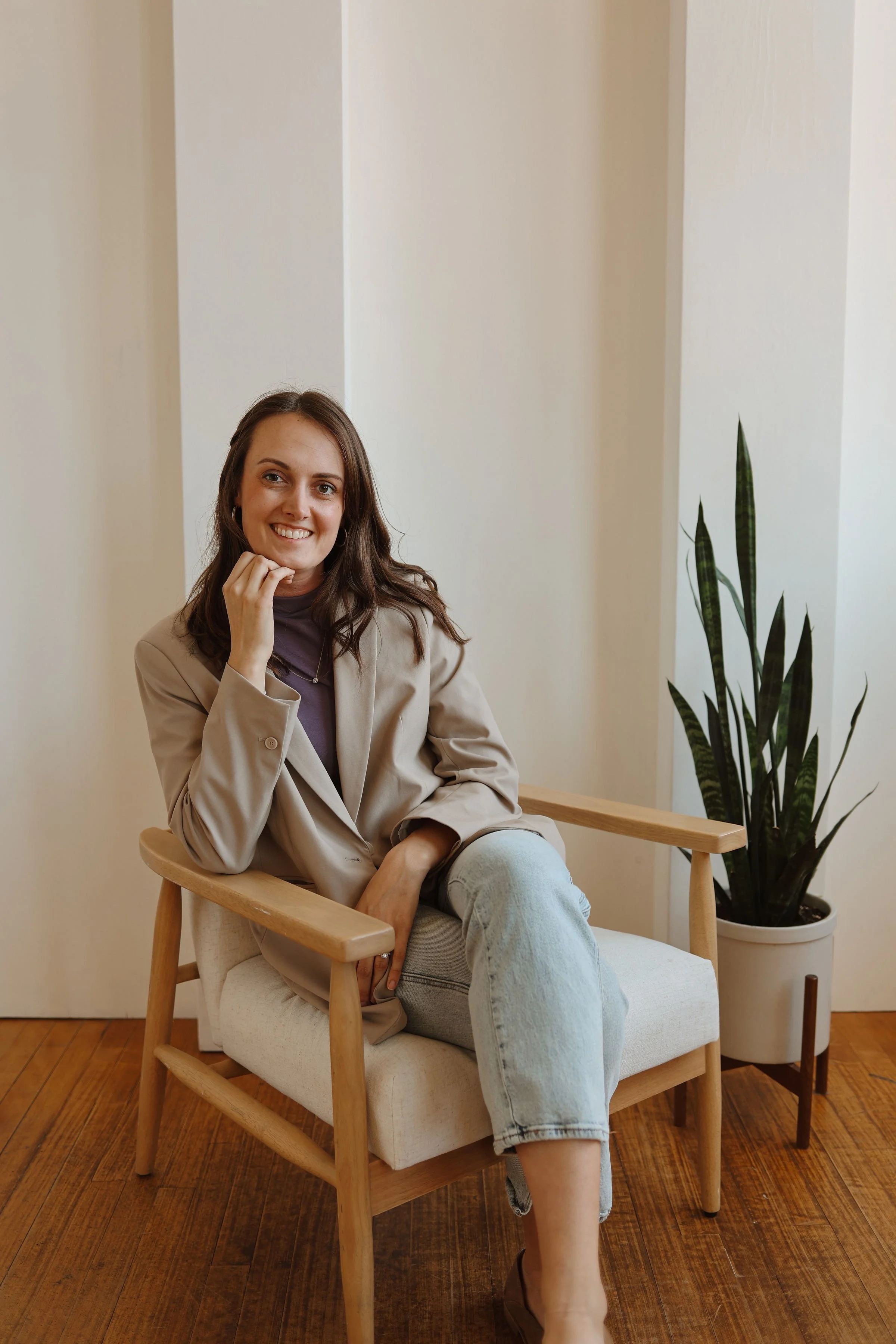 A young woman with brown hair and a warm smile sitting on a wooden chair with a cushion, wearing a beige blazer, lavender top, and light jeans, with a potted plant nearby and a plain light-colored wall in the background.