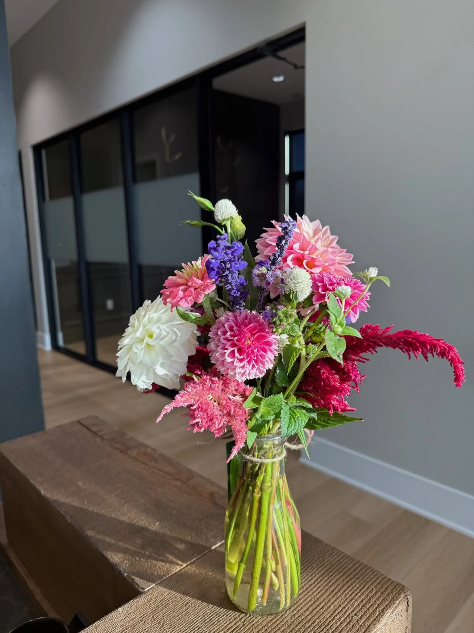 A bouquet of pink, purple, white, and red flowers in a clear glass vase on a wooden table in a modern indoor setting.