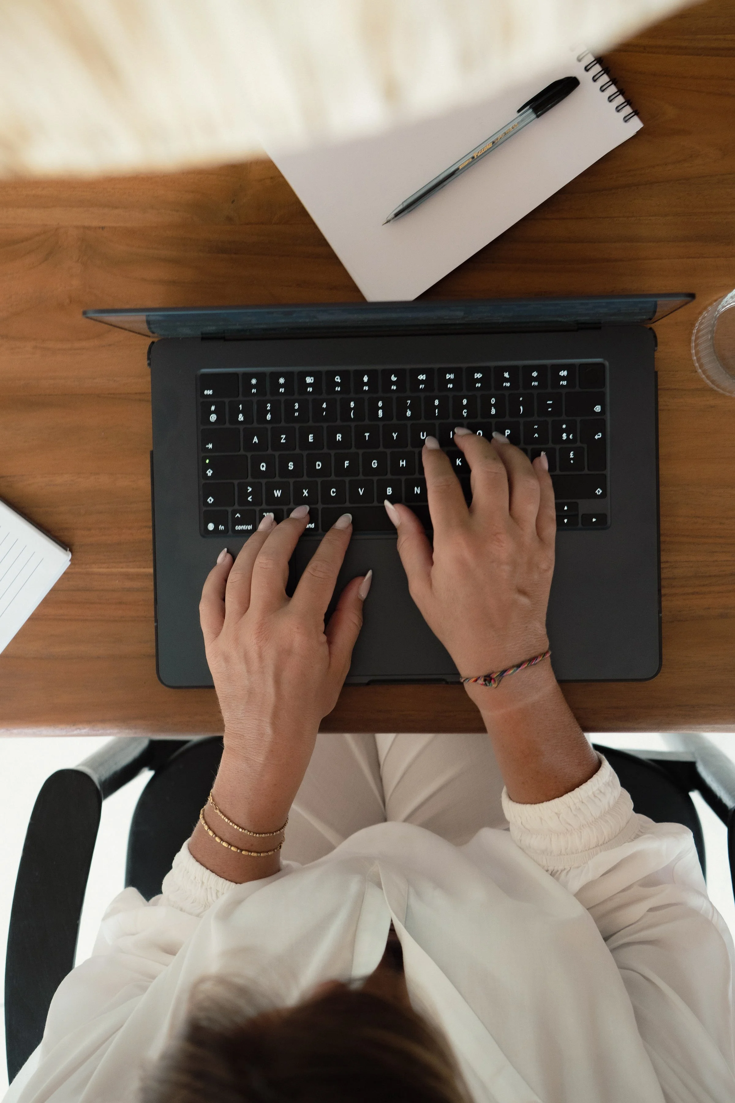 Overhead view of person typing on a laptop at a wooden desk, with a white notebook and a pen nearby.