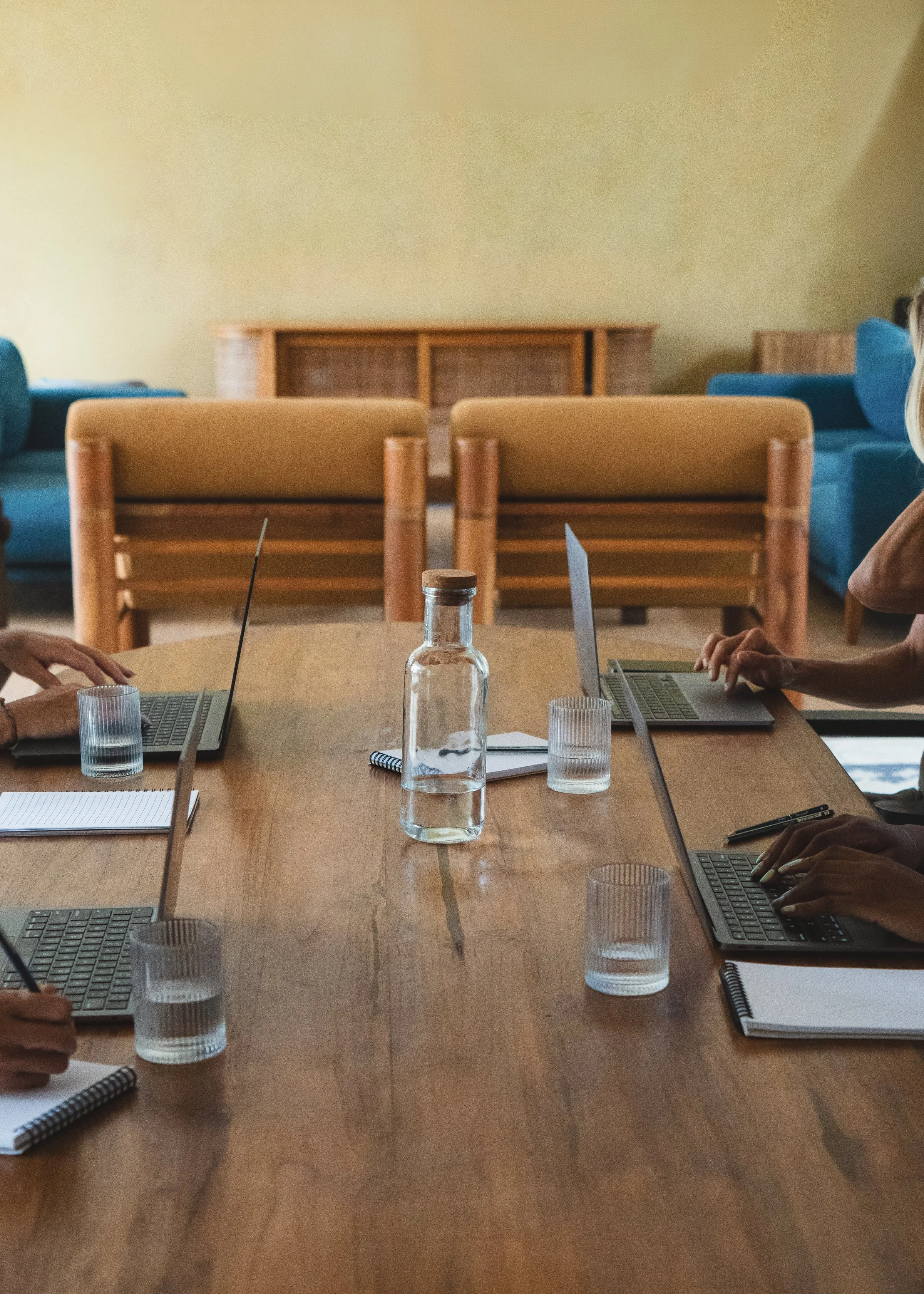 Meeting room with a wooden table, laptops, notebooks, glasses of water, and hands of people working.