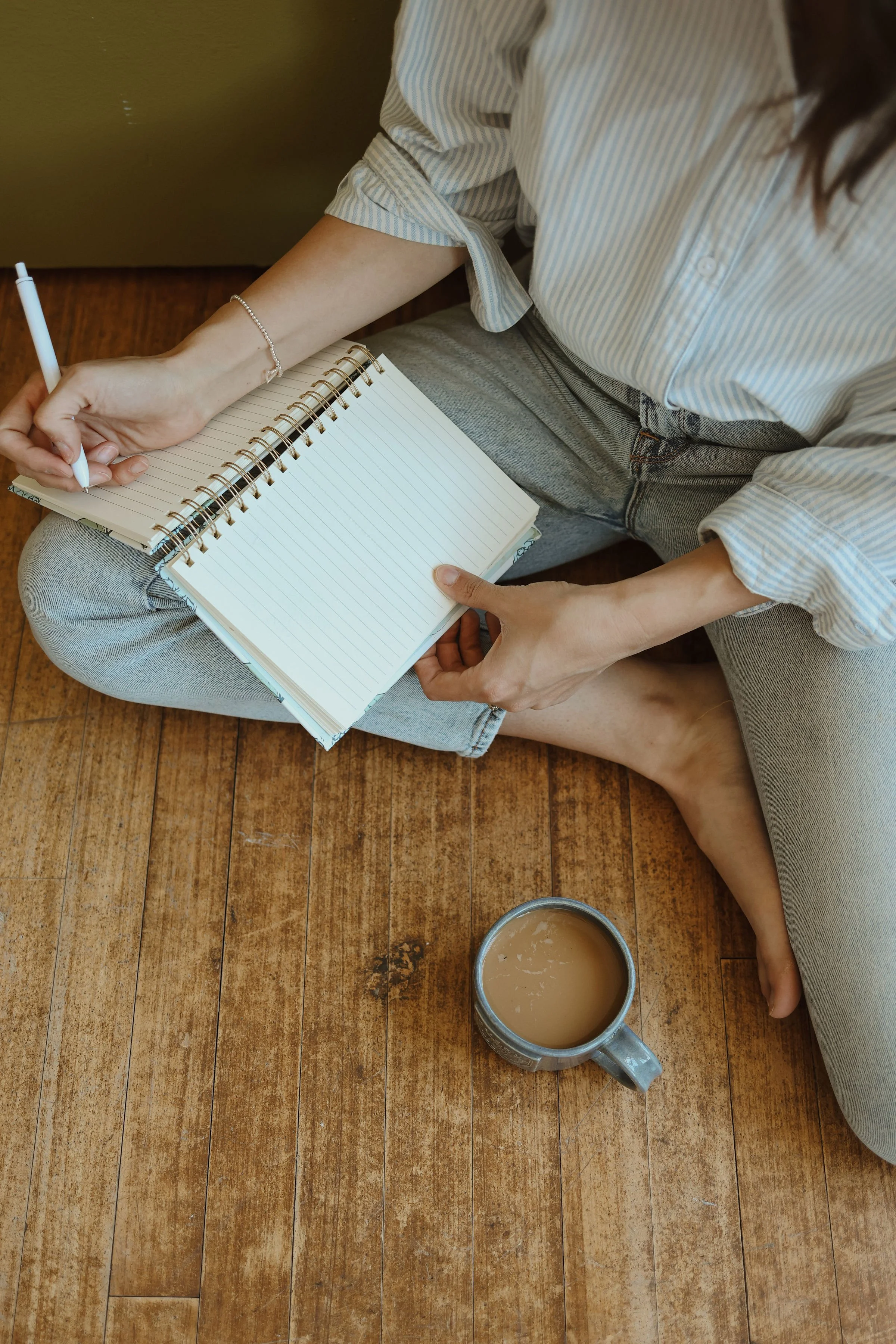 Person sitting on a wooden floor with a notebook and pen, a cup of coffee, and wearing a striped shirt and jeans.