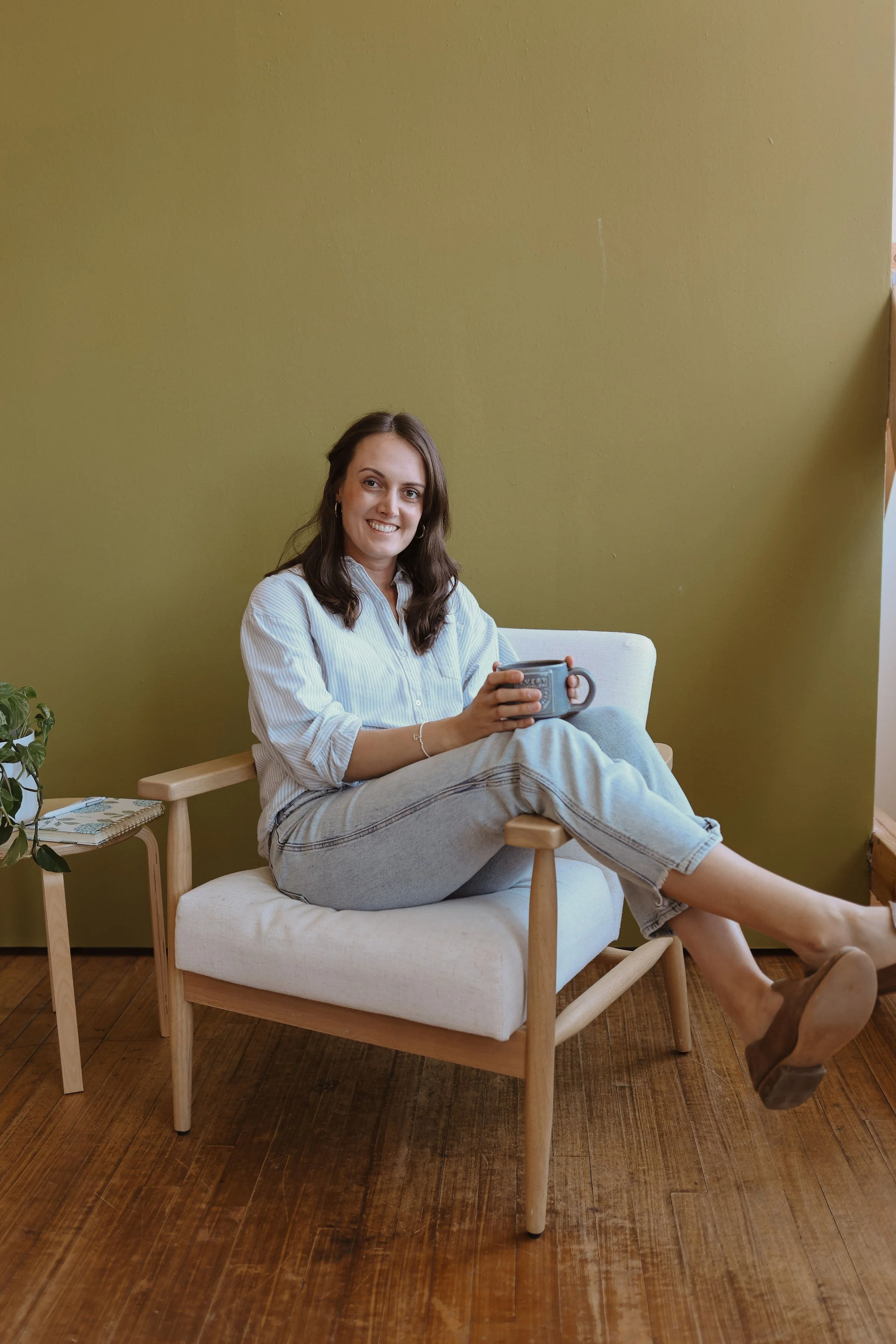 Marquel Stuedemann, a Christian Author and Speaker, sitting on a light-colored armchair with wooden legs, holding a mug, in a room with a green wall, wooden floor, and a small side table with a plant and a notebook.