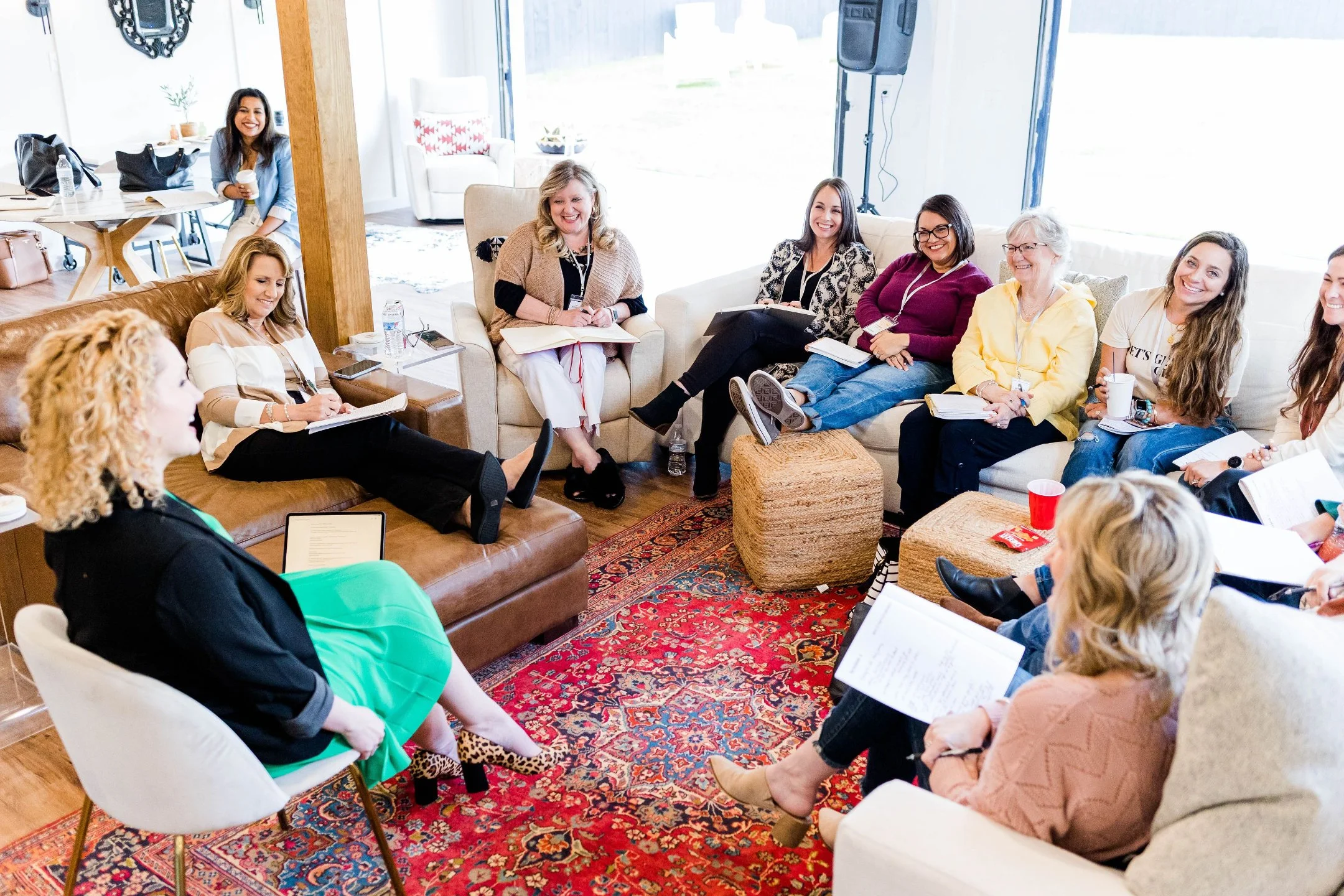 Group of women sitting and smiling during a meeting or workshop in a bright, casual space, with notebooks and drinks on their laps and around them.