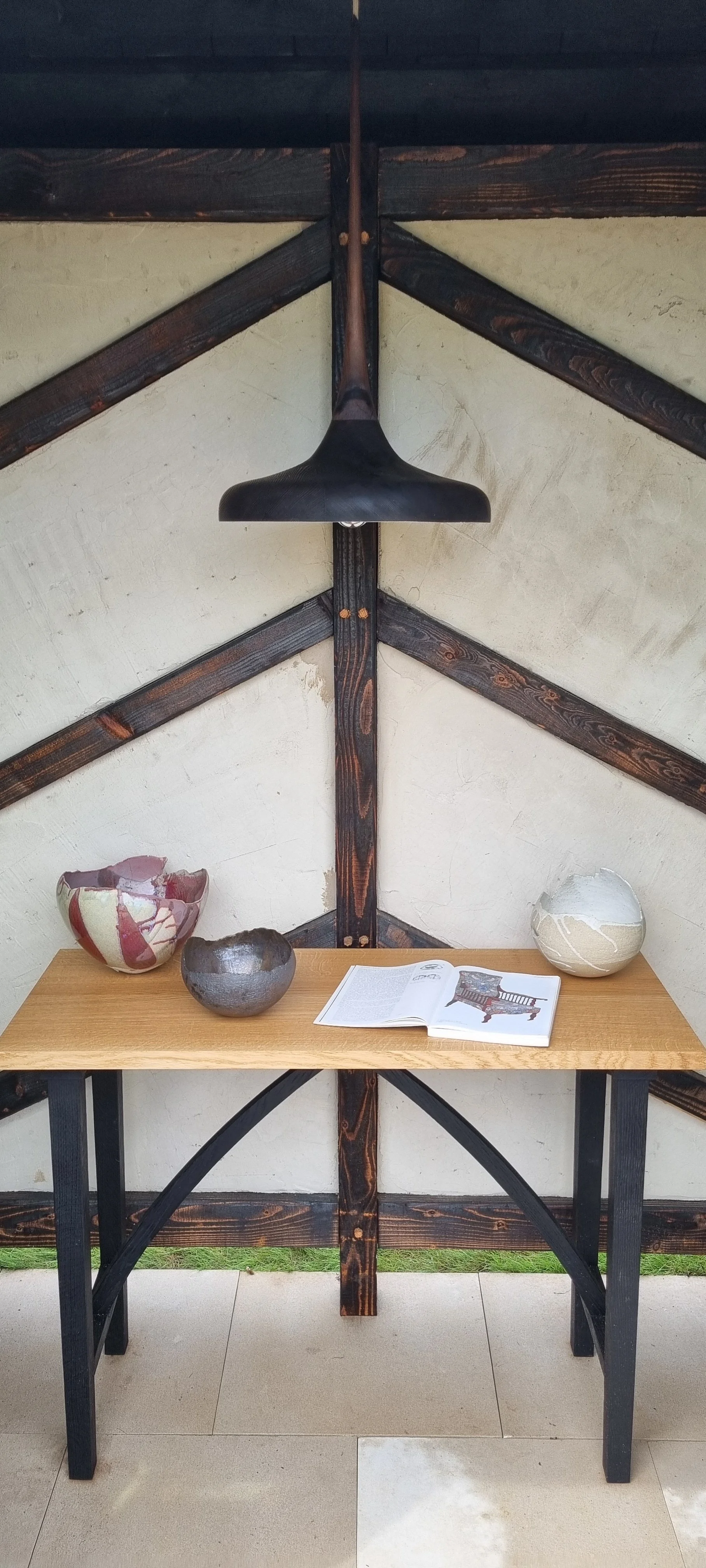 Decorative display with three ceramic bowls on a wooden table, an open magazine, and a black overhead light fixture against a rustic wall with dark wood beams.
