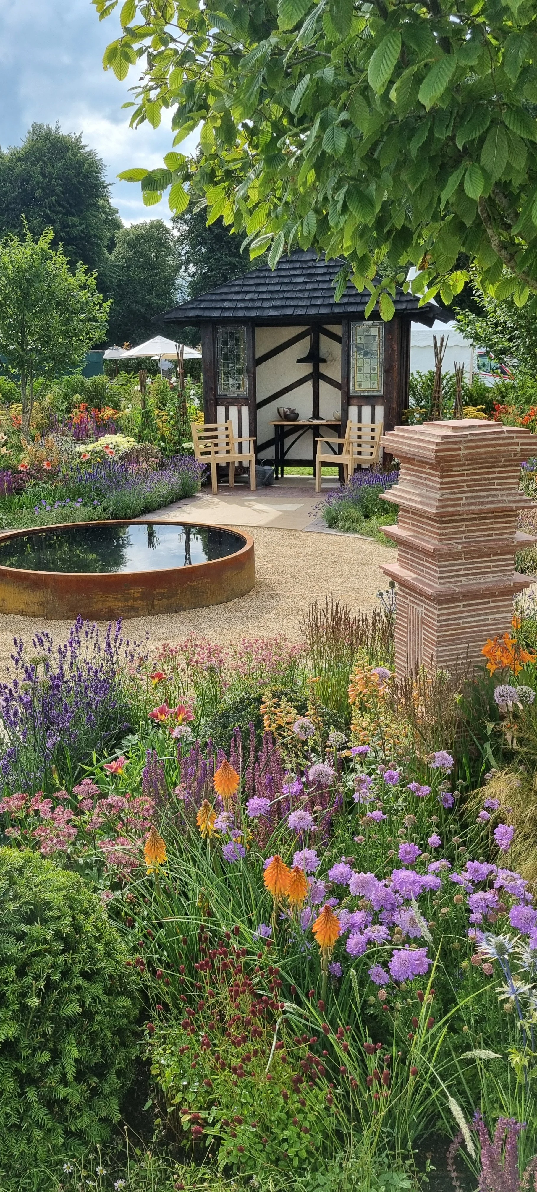 A garden with colorful flowers, a small pond, and a wooden gazebo with two chairs.