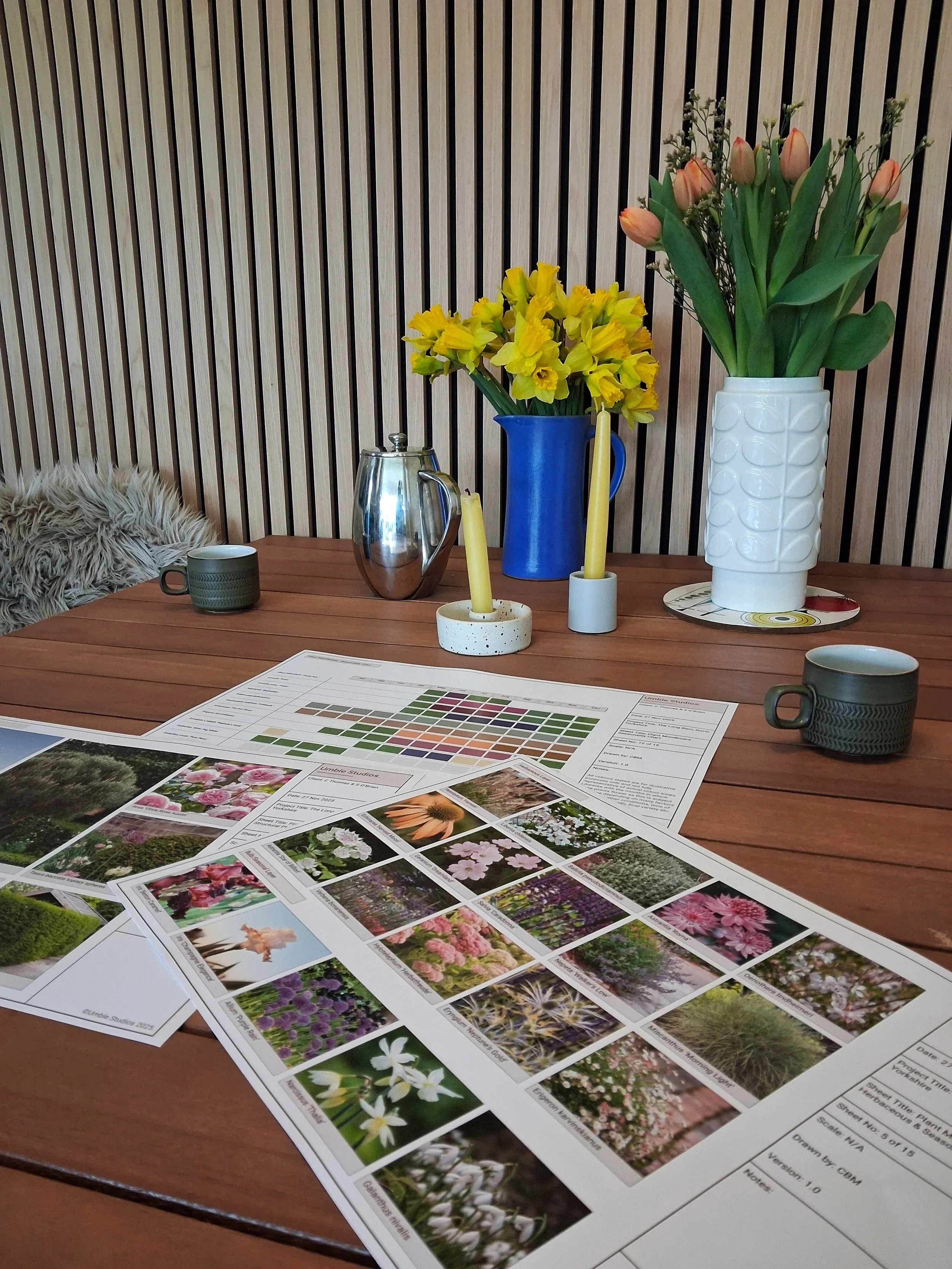 Design plan on a wooden table with color charts and flower photographs, two vases with yellow and peach flowers, candles, and coffee cups in front of a wooden slat wall.