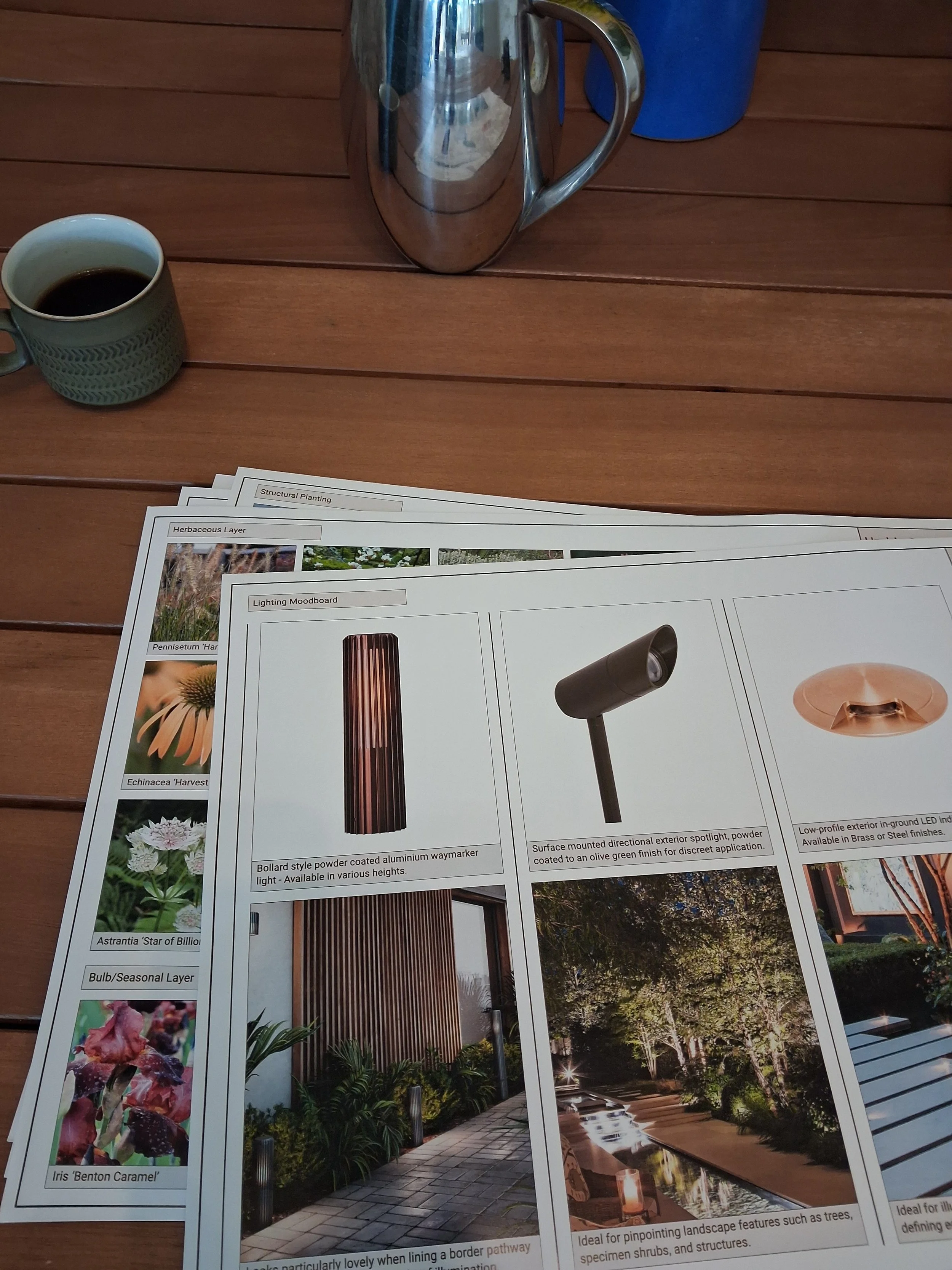 Outdoor wooden table with a cup of black coffee, a metallic pitcher, a blue cup, and several sheets of paper containing images and descriptions of landscape design elements.