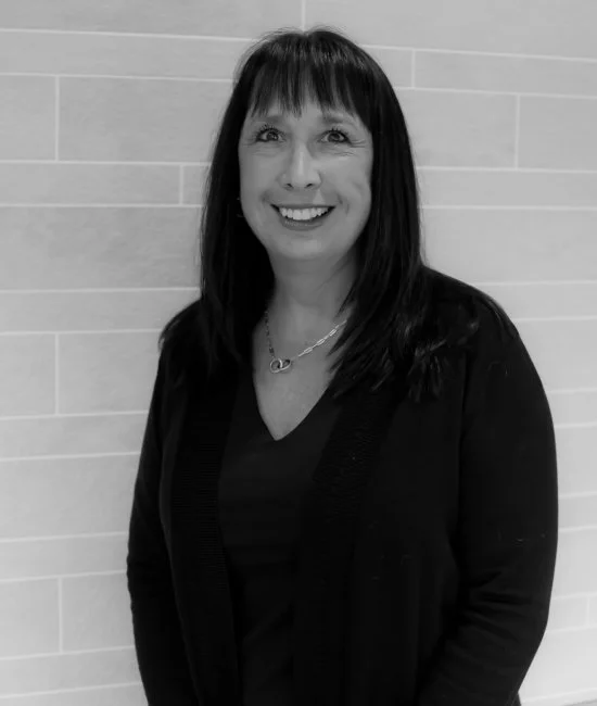 A smiling woman with shoulder-length dark hair and bangs, wearing a black jacket and a necklace, standing against a tiled wall.