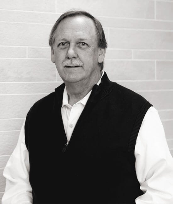 Black and white photo of an older man with light-colored hair and mustache, wearing a white shirt and a dark vest, against a tiled wall background.
