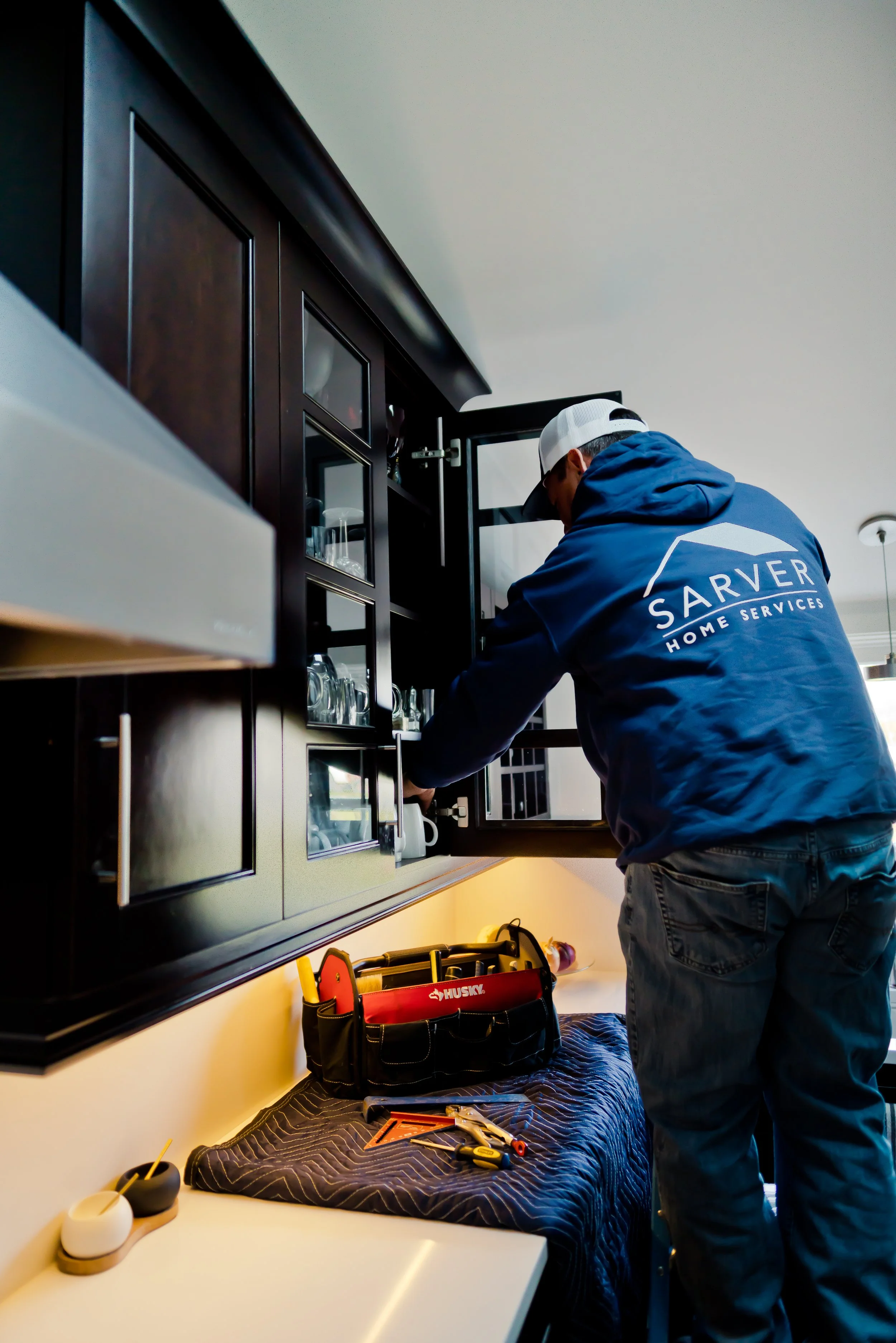 A person wearing a blue hoodie with 'SARVER HOME SERVICES' on the back, a white cap, and glasses, working on a kitchen cabinet door with tools on a table below.