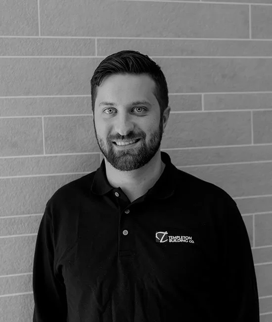 A man with dark hair and a beard, wearing a black polo shirt with a logo, standing in front of a brick wall, smiling at the camera.