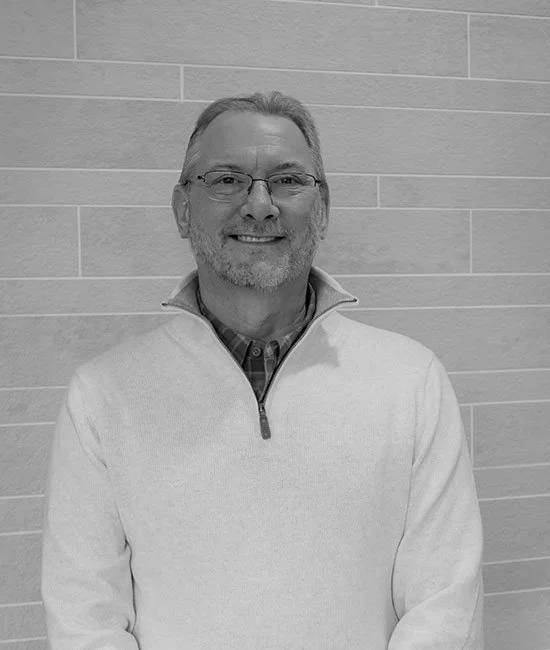 A smiling middle-aged man with glasses and gray hair, wearing a zip-up sweater over a shirt, standing in front of a light-colored tiled wall.