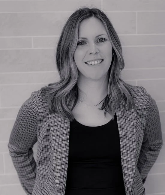 Woman with shoulder-length hair smiling, wearing a checkered blazer over a black top, standing in front of a brick wall.