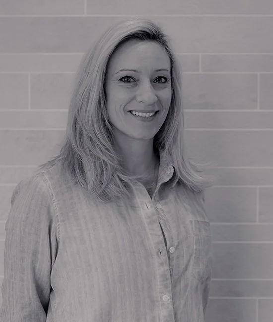 A woman with blonde hair smiling, standing in front of a tiled wall, wearing a light-colored button-up shirt.