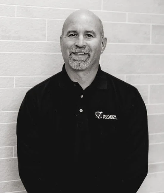 A middle-aged man with a beard and bald head, wearing a black polo shirt with a logo that says 'Templeton Building Co.', standing in front of a light-colored brick wall.