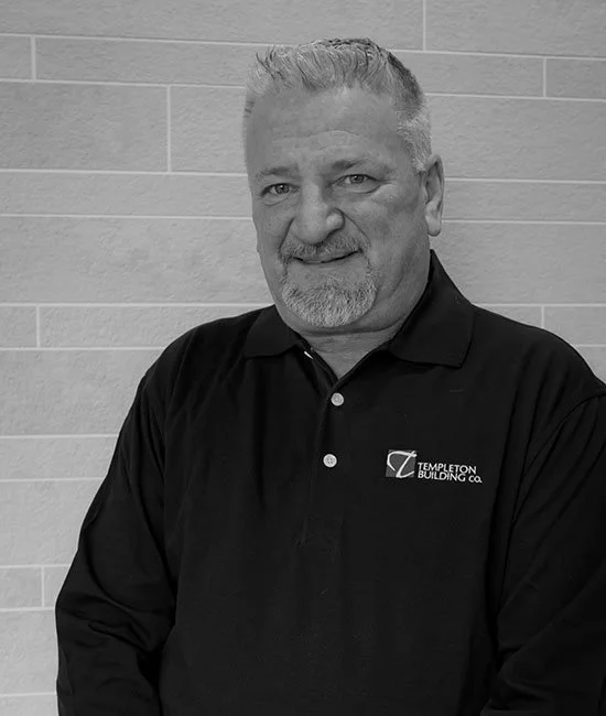 A middle-aged man with gray hair and beard, wearing a black polo shirt with a logo and text that reads "Templeton Building Co." standing in front of a tiled wall.